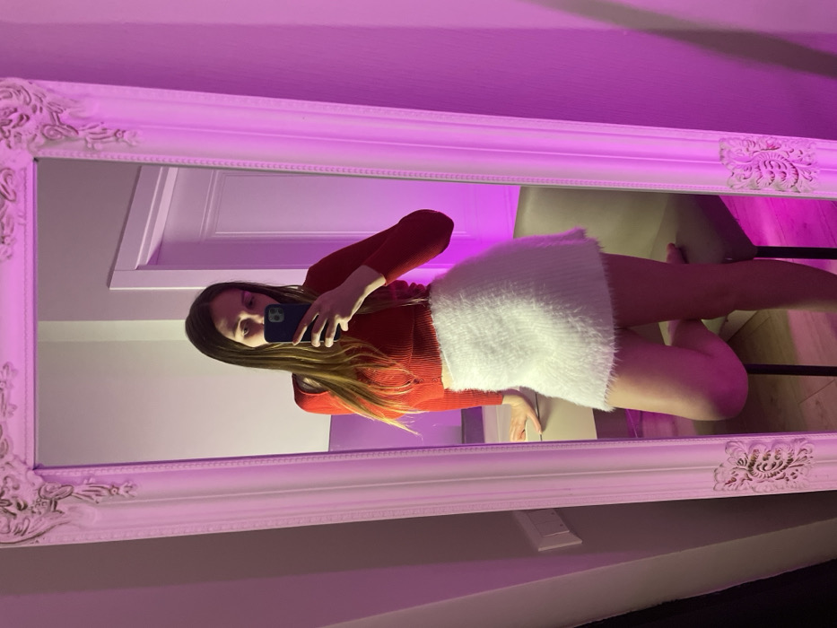 A young woman takes a mirror selfie wearing a red long-sleeved top and a white fluffy skirt. She holds her phone in her left hand. The mirror is ornate with a white frame and the background includes a white wall and a chair. The lighting appears to be purple, casting a vibrant hue on the scene.
