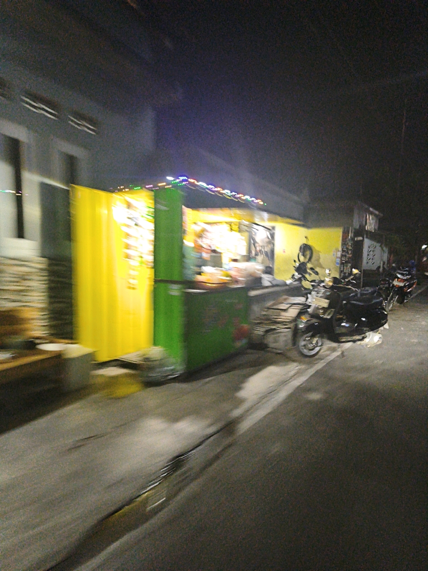 In Desa Pandeyan, Indonesia, at night, a blurry, low-light scene depicts a street-side food stall with a bright yellow and green exterior. Various items are visible within and around the stall, including what appears to be food preparation equipment and possibly packaged goods. In front of the stall, a black scooter is parked, and another motorcycle is visible further down the street. The ground in front of the stall is wet, with puddles reflecting the surrounding lights. The overall impression is of a busy, possibly late-night food vendor operating in a common urban or semi-urban Indonesian setting.