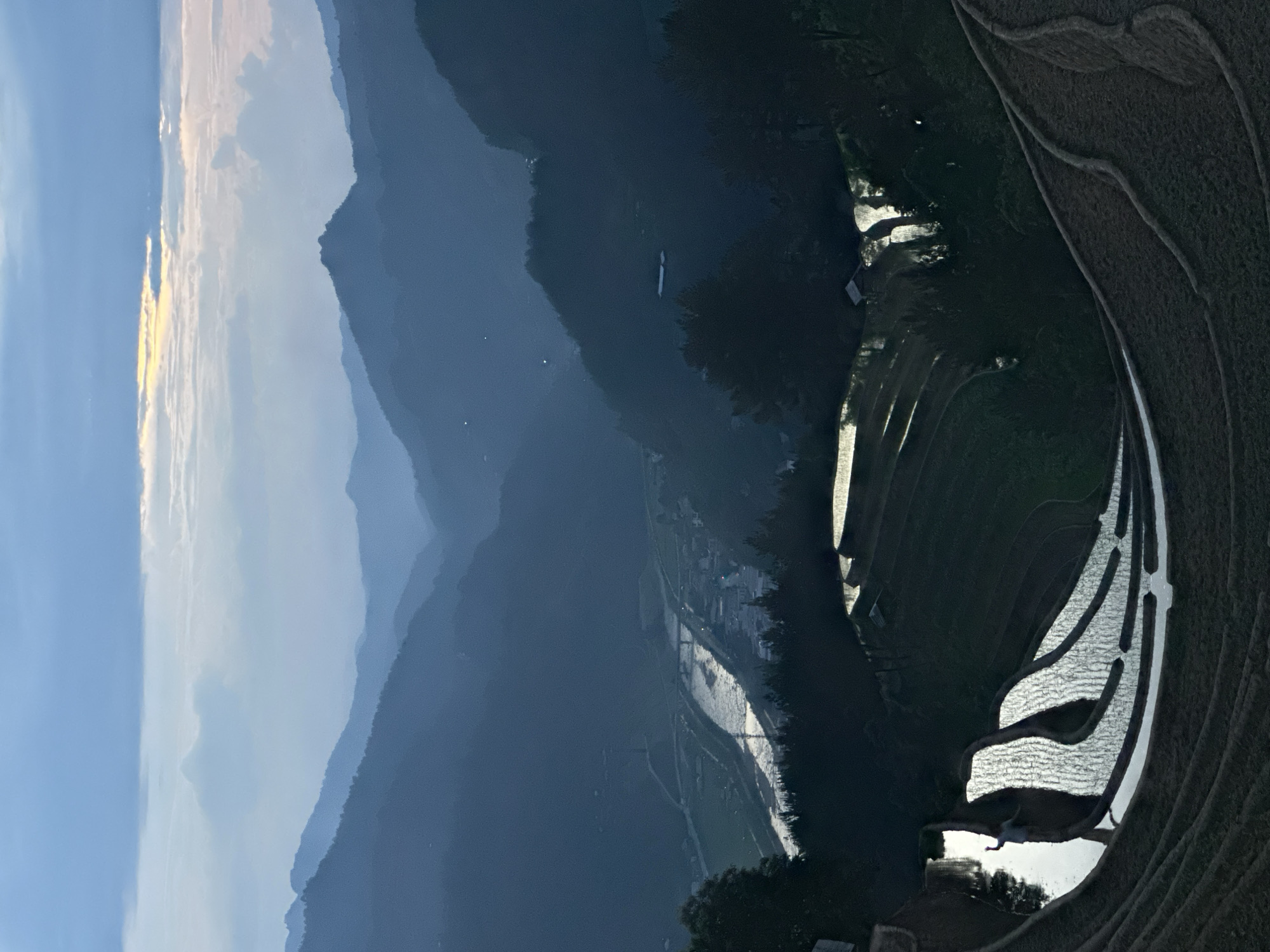 This panoramic image captures a breathtaking landscape in Mù Cang Chải, Vietnam, at either sunrise or sunset. The sky, a gradient of light blue, transitions to soft yellow and orange hues along the horizon, where scattered clouds are illuminated by the low sun.
In the background, multiple layers of mountains recede into the distance, appearing increasingly hazy and desaturated due to atmospheric perspective, creating a sense of depth. The closer mountains are dark and silhouetted against the brighter sky.
The midground reveals a winding valley, where a river or stream snakes through, reflecting the sky's light as a bright, silvery ribbon. Small, indistinct lights or structures dot the valley floor, hinting at a remote village or settlement nestled among the hills.
The foreground and right side of the image are dominated by the intricate patterns of terraced rice fields. Many of these terraces are filled with water, which brilliantly reflects the bright sky, creating shimmering, curvilinear bands of light against the darker, un-submerged earth. The geometric yet organic shapes of the terraces are a testament to human cultivation within a grand natural setting.
The overall lighting creates stark contrasts between the illuminated water and sky and the shadowed mountains and land, emphasizing the dramatic topography. No people or visible text are discernible in the scene. The image conveys a serene and majestic atmosphere, showcasing the iconic beauty of Vietnam's terraced landscapes.
