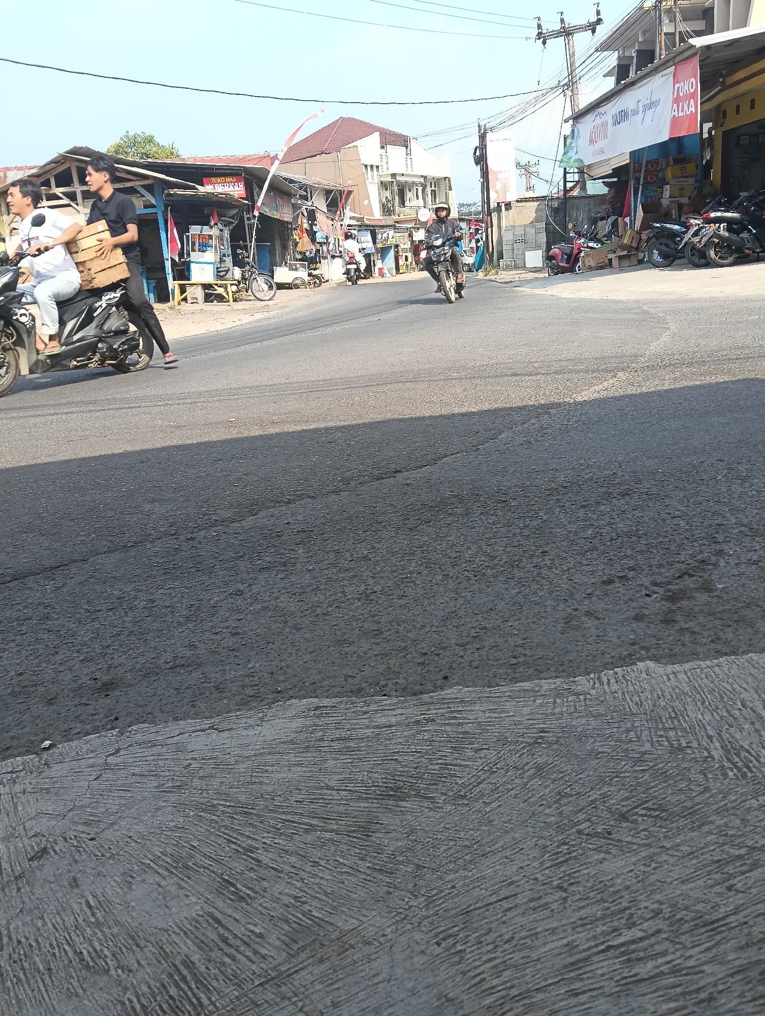 The image shows a street scene in Puspahiang, Indonesia. Two young men are riding a motorcycle, with the passenger carrying a stack of wooden planks. The motorcycle is dark-colored and appears to be a scooter. They are on a paved road with a shadow cast across it from a nearby building. To the right of the road, there are several small shops with awnings, one of which has a sign that says "TOKO ALKA". A few other motorcycles are parked in front of the shops. The buildings appear to be low-rise structures typical of a commercial area in a town. The sky is clear and blue, suggesting a sunny day. Electrical wires are visible overhead, supported by utility poles. The overall scene depicts everyday life and commerce in a local Indonesian community.