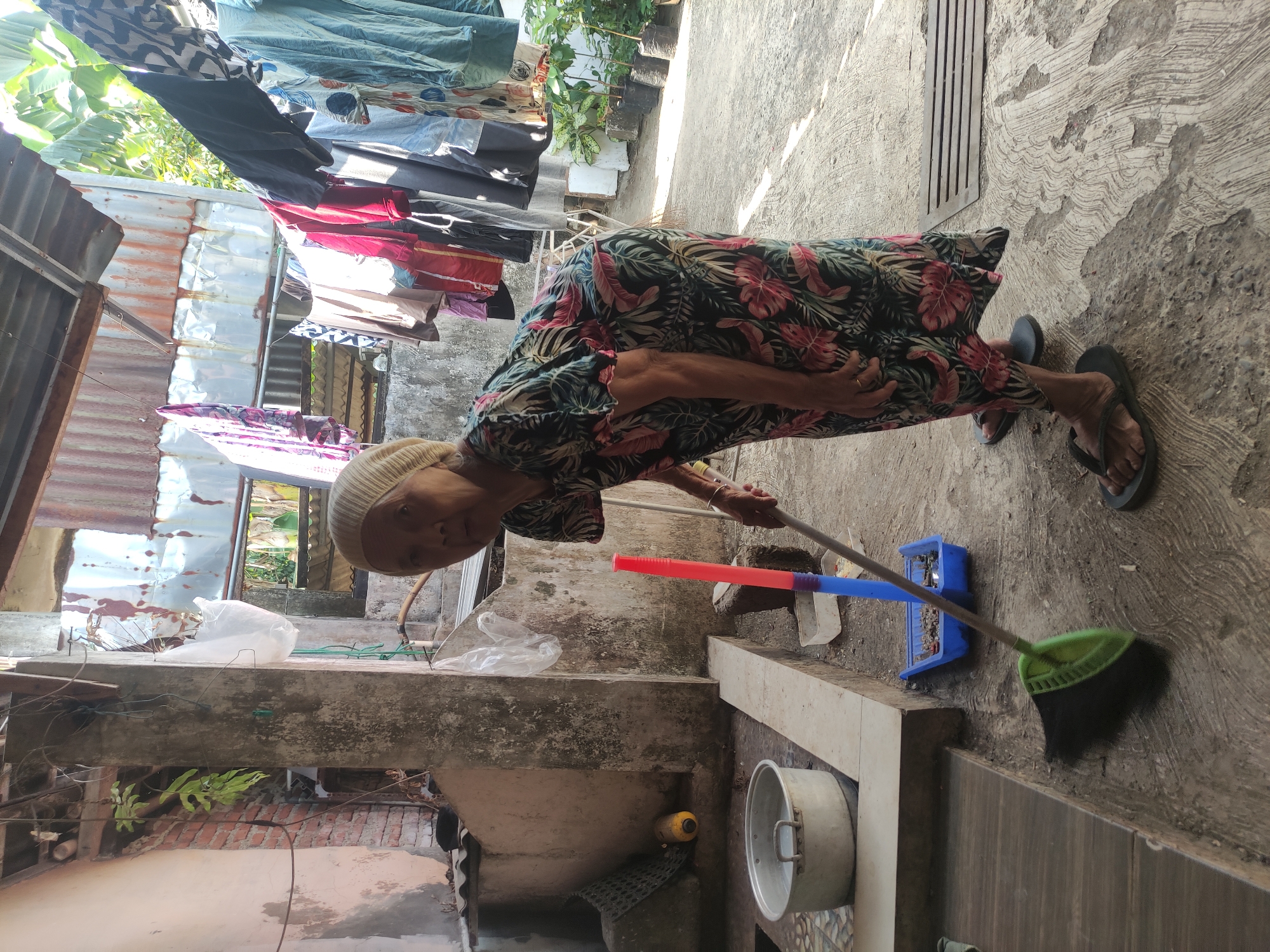 In Klampok, Indonesia, an elderly woman, wearing a headscarf and a floral-patterned dress, is sweeping a concrete outdoor area with a green-bristled broom. She is bent over, holding the broom and looking towards the camera. A blue dustpan with a red and blue pole stands beside her. Nearby, a large metal pot sits on a raised platform. Clothes are hanging on a clothesline in the background, above potted plants. The environment appears to be a residential yard, with elements of weathered construction and lush vegetation.