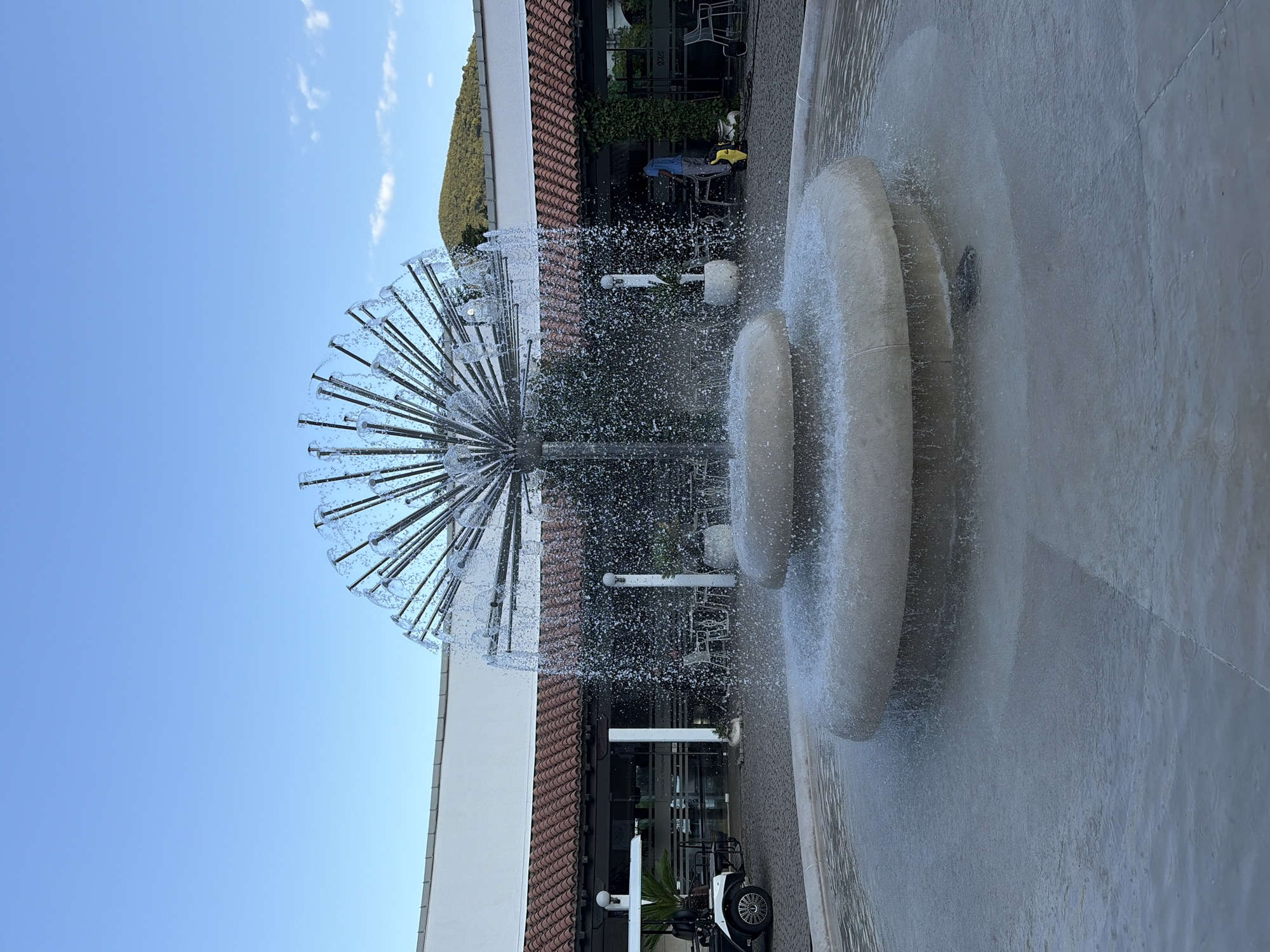 An outdoor fountain is featured in the foreground, with water spraying from a metal structure resembling a dandelion seed head. The fountain has two circular tiers. In the background, a building with a white facade and a red-tiled roof is visible. To the left of the building, a white golf cart is parked. To the right, a person in a blue shirt and gray pants is operating a yellow cleaning device near some outdoor seating and tables. A green, tree-covered hill is visible in the distance under a clear blue sky with a few wispy clouds. The scene appears to be in a public area, possibly a resort or a plaza, with paved ground around the fountain. The image is taken during daylight.