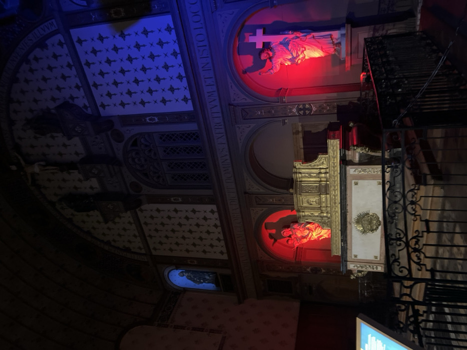 The image depicts an interior view of a historic hall, specifically identified as a section of the Musée des Beaux-Arts de Dijon, France, likely within the former chapel of the Palace of the Dukes of Burgundy. The scene is dimly lit, with strategic colored illumination highlighting various architectural and sculptural elements.
The hall features a high arched ceiling and walls adorned with patterned motifs, appearing as dark cross-like symbols against a lighter background. Decorative moldings, pilasters, and arched recesses structure the space. A large arched window with a grid pattern is visible in the upper central section. A Latin inscription, partially legible as "NOMINAS GLORIA DICTAE DOMINUS EXERCITVVM ET IN ISTO LOCO," runs along a frieze above the lower architectural elements.
At the lower level, a gilded, ornate, multi-tiered retable or altar-like structure rests on a marble base with a central golden medallion. This structure is flanked by two arched niches, both illuminated in red light. The left niche contains a sculpture of a Madonna and Child. The right niche houses a sculpture of a male figure with an uplifted arm, standing alongside a white cross.
In an upper arched niche on the left, a dark sculpture of a robed figure is visible, illuminated by blue light. A crucifix is mounted on the wall above the central window. The upper right section of the wall is also washed in blue light, emphasizing its patterned design.
In the foreground, a black metal railing with scrollwork separates the viewing area from the display. A partially visible, modern illuminated display panel, showing blue text on a dark background, is present on the far left. No individuals are present in the scene.