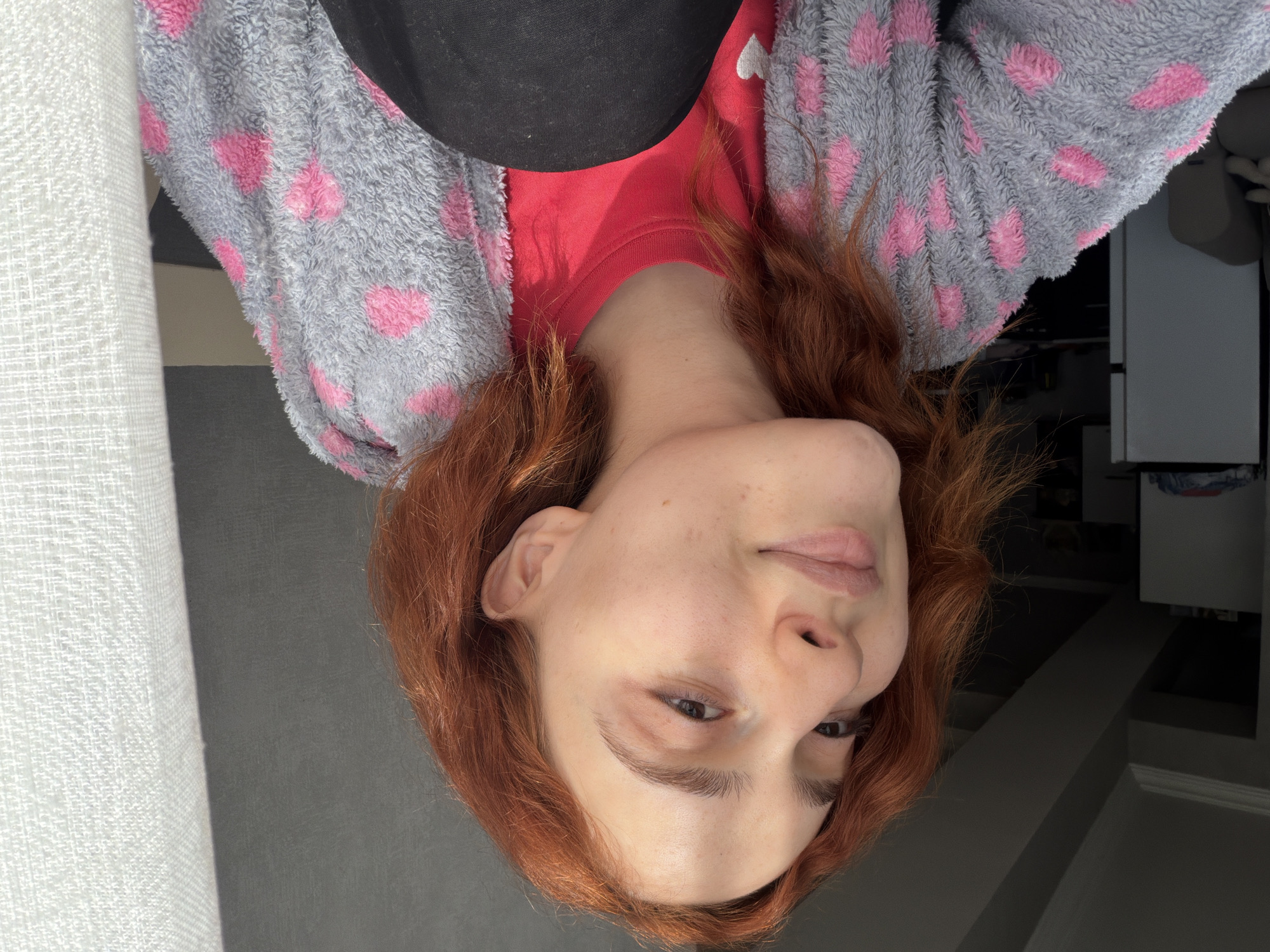 A female-presenting individual with fair skin and red hair is positioned at an angle in the frame. She is wearing a red shirt, covered by a grey, fuzzy robe with pink heart-shaped patterns. A black object, potentially a piece of clothing or accessory, is visible above the shirt. The background includes a white textured curtain or fabric on the left side and a grey wall or surface beneath her face. In the upper right corner, there is a glimpse of furniture or a cabinet with a light-colored door or drawer. Without additional context or reference points, it is impossible to determine the location in Tbilisi, Georgia.