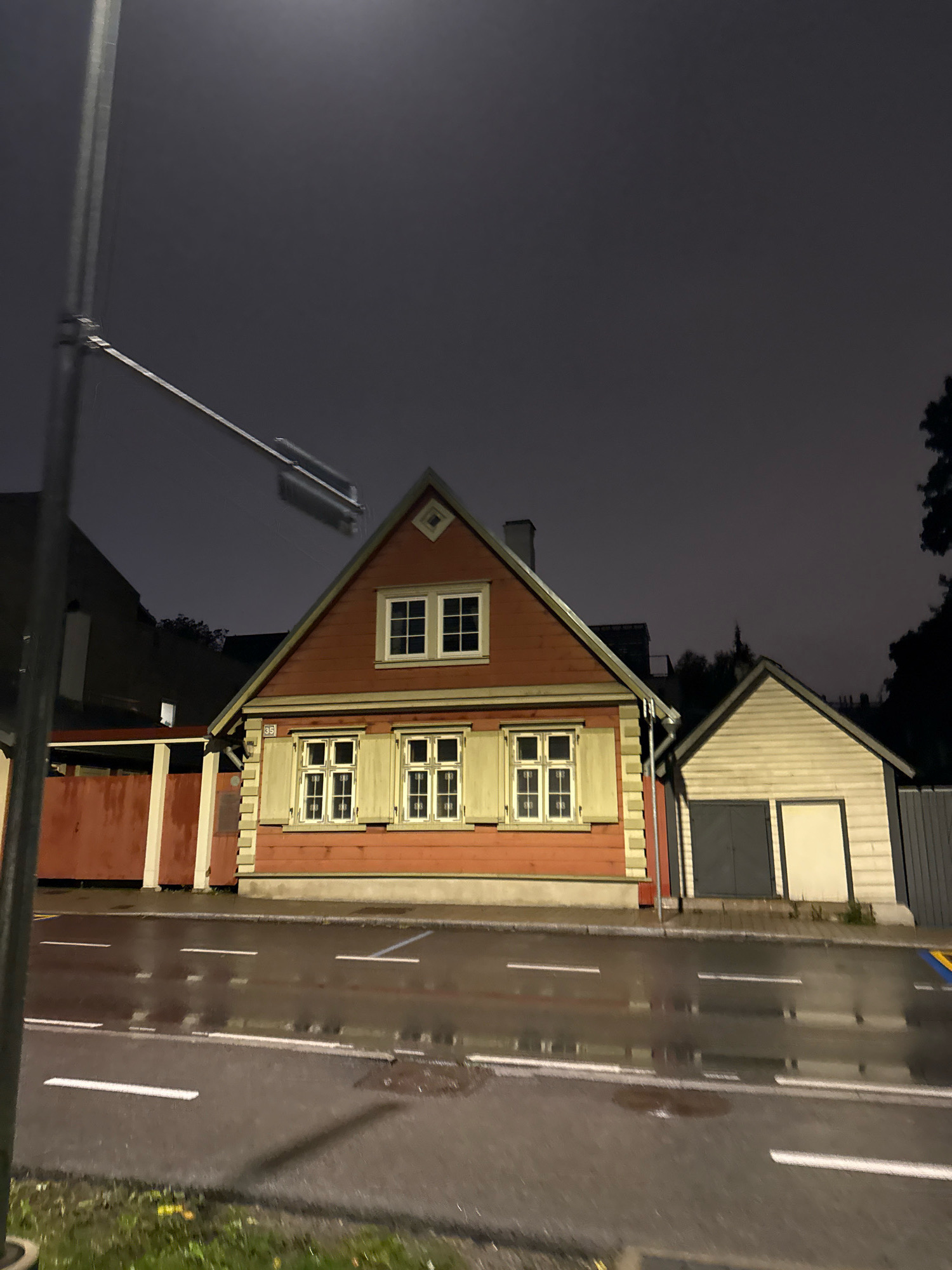In Tallinn, Estonia, on a dark, overcast night, a wet road with white lane markings reflects the dim light. To the right of the road, a sidewalk leads to a collection of buildings. The primary structure is a two-story wooden house painted red with white trim, featuring a prominent gable roof. It has several windows with light-colored shutters. Adjacent to this house is a smaller, single-story wooden outbuilding, possibly a garage, painted beige with dark gray double doors and a lighter beige rectangular panel. The scene is partially illuminated by a street light on the left.