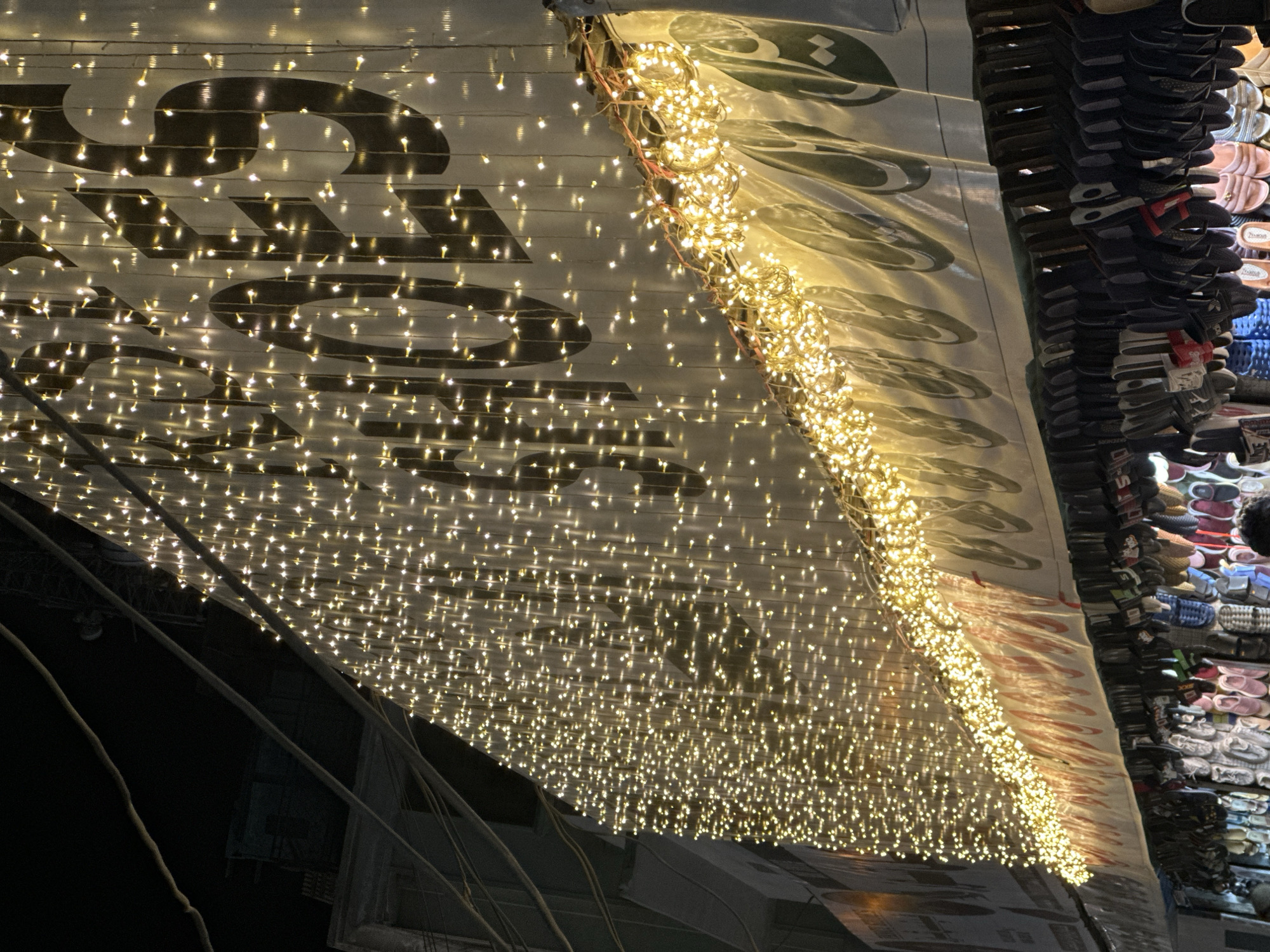 Captured at night in Quetta City Tehsil, Pakistan, the image shows a low-angle view of a commercial storefront awning draped with strands of tiny, warm-toned string lights. The awning is white with large, stylized black text partially visible through the dense mesh of lights. To the right, a vertical display of shoes, primarily sandals and flip-flops in various colors and sizes, is visible. The background contains blurred structural elements of the surrounding buildings.