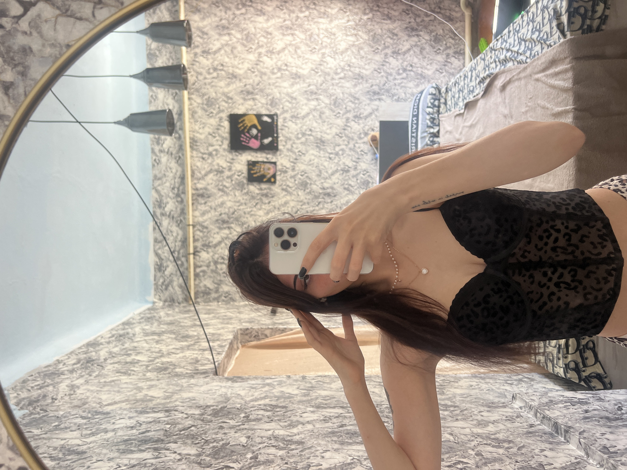 A young woman with long dark hair is taking a selfie in a mirror. She is wearing a black leopard-print corset and a pearl necklace. Her left hand is raised to her head, and her right hand is holding a smartphone. The background of the image appears to be a bedroom with textured wallpaper, a light blue ceiling, and three pendant lamps. A bed with patterned bedding is visible in the background. The image was taken indoors.