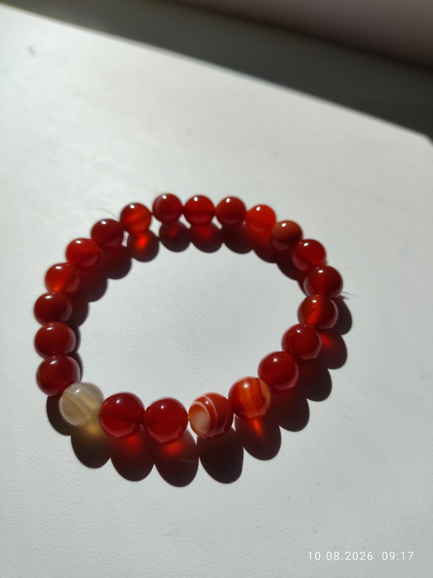 A close-up, high-angle shot shows a red beaded bracelet lying on a white, textured surface. The bracelet is arranged in a circular shape, with some beads casting shadows. The lighting creates a dramatic effect, with bright highlights on the polished beads and deep shadows. The beads themselves are predominantly a deep, translucent red, with a few exceptions. There are two beads towards the bottom left that appear lighter, almost translucent yellow or white, with some striations. Two beads further along in the circle also exhibit prominent banding in shades of red and white. The surface the bracelet rests on has a subtle texture, possibly paper or a similar material. A dark, out-of-focus area is visible in the upper part of the image, suggesting an overhead object or shadow. In the bottom right corner, a timestamp is visible: "10.08.2026 09:17". The scene does not contain any identifiable elements that would place it in the city of Pavlodar, Kazakhstan.