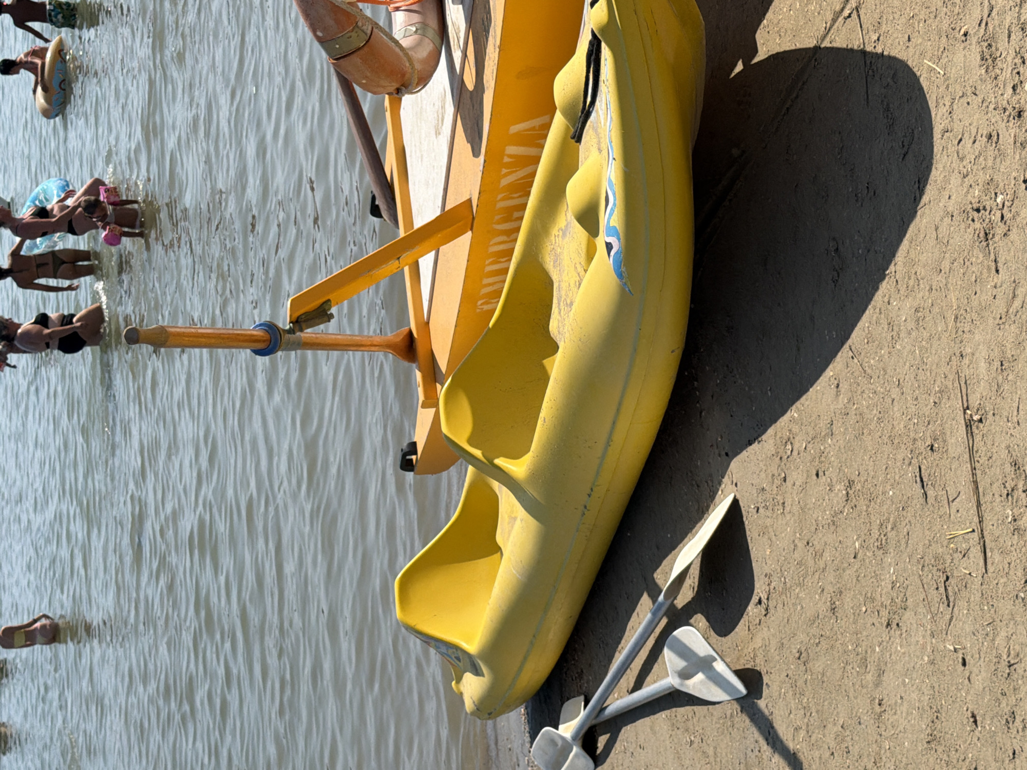 A yellow pedal boat is beached on a sandy shore beside the sea in Gatteo, Italy. Two paddles, one white and one with a grey shaft and white blade, lie in front of the pedal boat. In the background, several people are in the water. One person is seated on an inflatable ring, while others stand or wade in the shallows. The sea is calm with gentle ripples. A bright orange raft with the word "EMERGENZA" is also visible behind the pedal boat. The sun casts a shadow from the pedal boat onto the sand.
