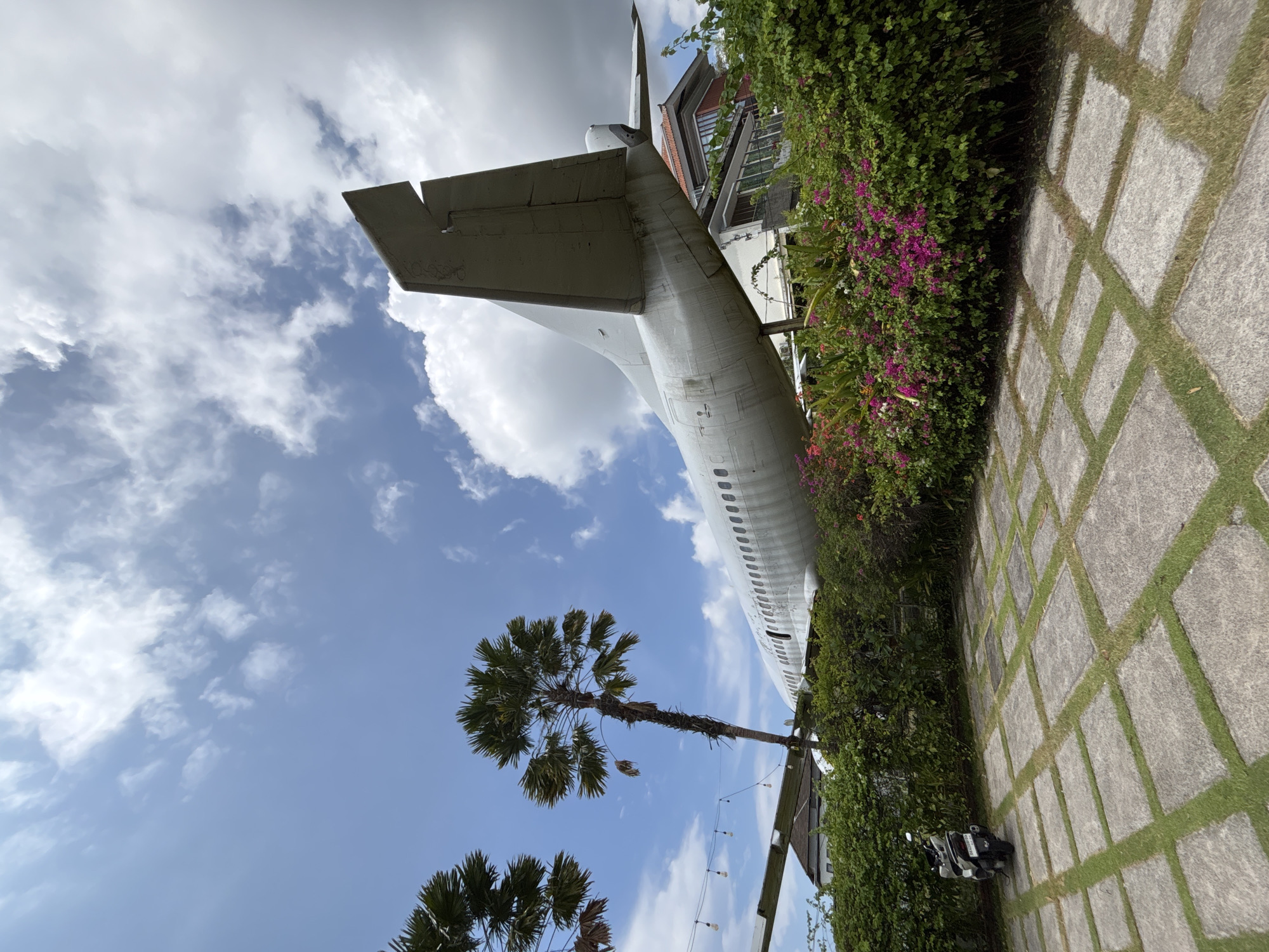 The image shows an airplane structure in Kuta, Indonesia. The tail and part of the fuselage of a white aircraft extend upwards from a garden area. The tail is plain with possible markings. The fuselage has a visible line of windows. A building with a multi-level structure is partially visible next to the airplane. The garden features lush greenery, including flowering bushes with purple blooms, alongside a grid of paved stones with grass growing between them. A palm tree is visible below, partially obscuring the sky with its foliage. The sky is blue with scattered white clouds. A scooter is parked below.
