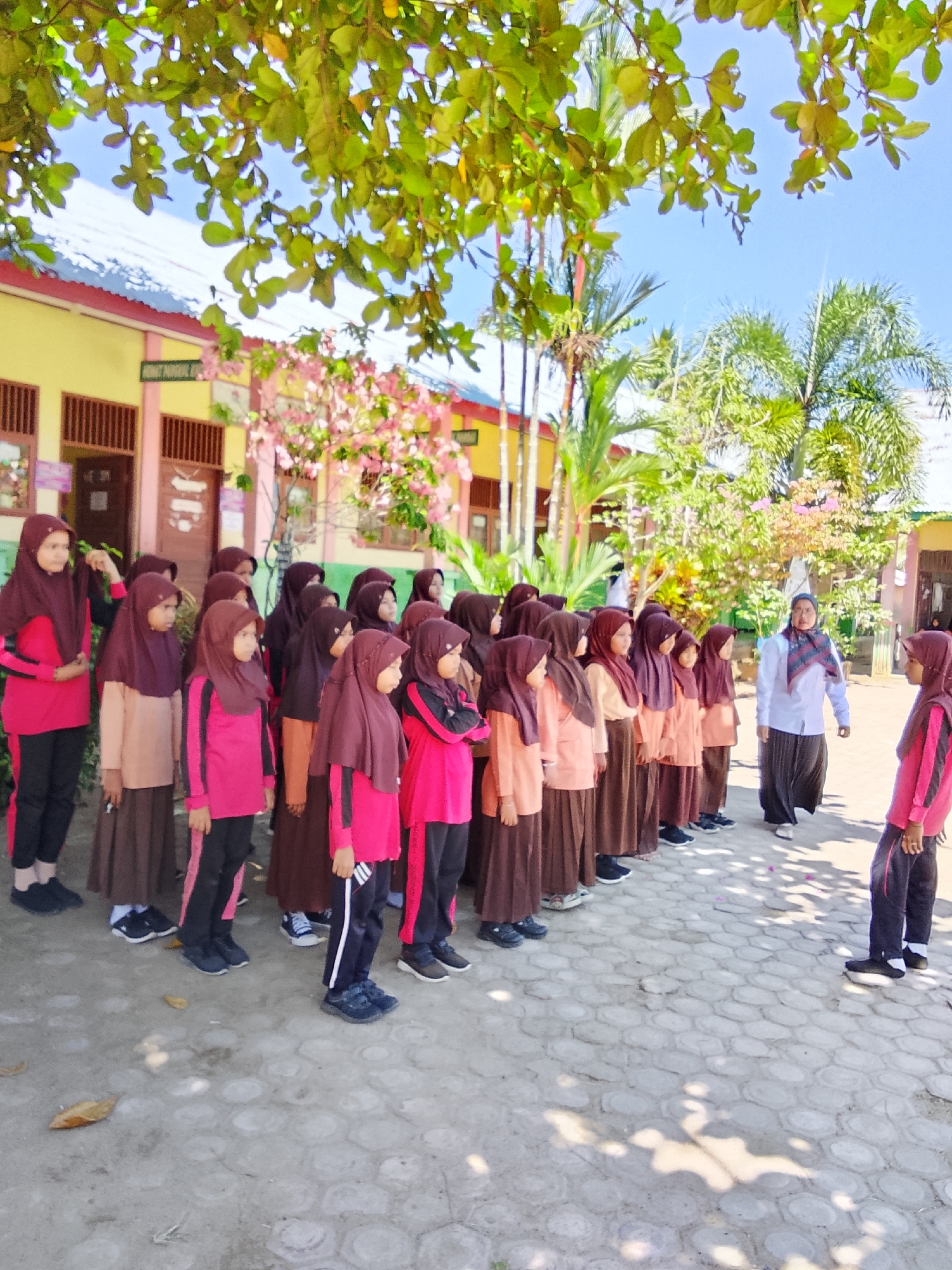 In Baratpayaitek, Indonesia, a group of female students stand in formation outdoors on a paved courtyard. The students, dressed in Islamic school uniforms consisting of long skirts and hijabs, are primarily in shades of brown and pink. Some students are looking towards a woman standing to their right, who is also dressed in a long skirt and a white blouse with a patterned scarf. The environment features a building with a yellow facade and a red roof in the background, along with trees and foliage. The ground is paved with interlocking hexagonal stones. The sky is clear and blue.