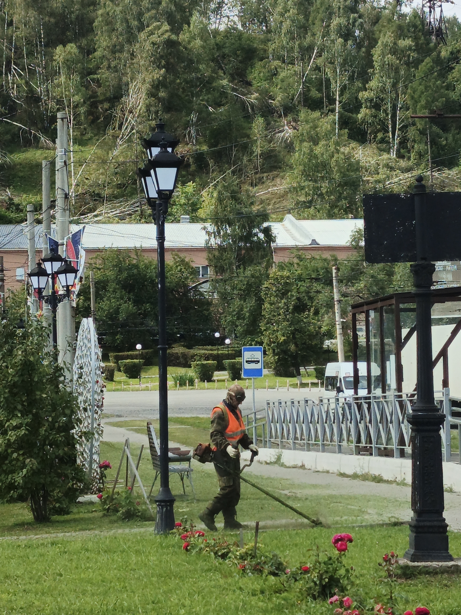A man in a green uniform and an orange reflective vest is using a string trimmer to mow a grassy area. He is wearing protective eyewear and gloves. Behind him, a black lamppost stands prominently. In the background, a white minibus is visible, along with a bus stop sign. The scene appears to be in a public park or along a street, with trees and bushes framing the area. The overall impression is one of routine maintenance work in an outdoor urban setting. The image is believed to be taken in Zlatoust, Russia.