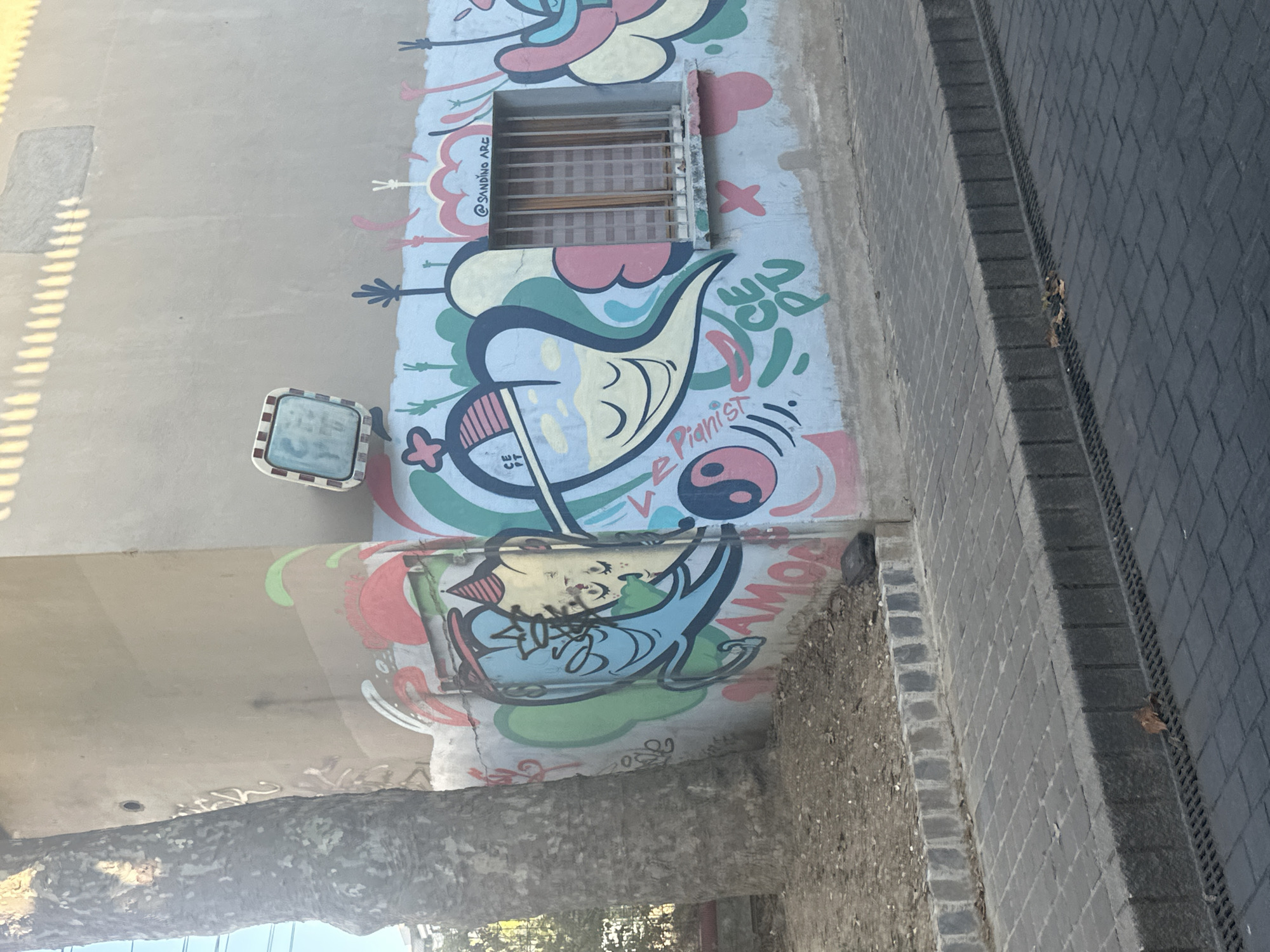 The image captures a section of an outdoor wall adorned with colorful graffiti. A large tree trunk is visible on the left, its rough bark contrasting with the smooth painted surface of the wall. The graffiti features cartoon-like figures, abstract shapes, and text. One prominent element is a large heart shape with a face. A barred window is integrated into the wall, also surrounded by graffiti. The wall itself appears to be part of a building, with a portion of its concrete facade visible above the graffiti. In the foreground, a sidewalk made of dark paving stones runs along the base of the wall, separated by a stone curb. A drain grate is also visible on the sidewalk. The environment suggests an urban setting, possibly a street or alleyway. The style and subject matter of the graffiti hint at an artistic and possibly youthful expression. The presence of a barred window might suggest the building is occupied or has some security features. The overall impression is one of a vibrant, urban art installation.