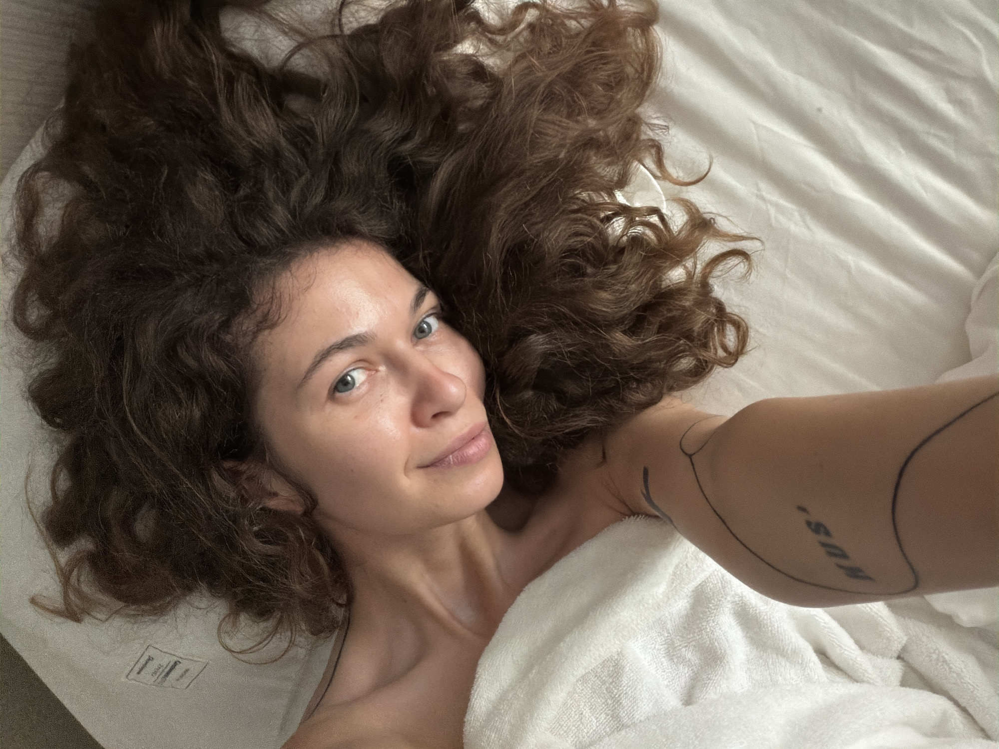 A woman with long, voluminous, dark brown curly hair and blue eyes lies on white bedding, taking a selfie from an overhead perspective. Her head is positioned towards the upper left of the frame, with her hair fanned out around her head and shoulders, covering much of the white sheets. She has a serene and slightly smiling expression, looking directly at the camera. A white towel or robe is draped over her body, covering her chest and right arm, implying she is partially clothed or wrapped up.
On her left bicep, a black line-art tattoo is visible, which appears to form abstract shapes and includes the partial text "NUS'". The bedding is light-colored, likely white, with subtle wrinkles. In the background on the left, a portion of a white label or tag is visible on what appears to be a mattress or pillow. The lighting is soft and even, suggesting an indoor setting during the daytime. The overall scene conveys a sense of comfort and relaxation, possibly in a hotel room or bedroom in Hà Nội, Vietnam.