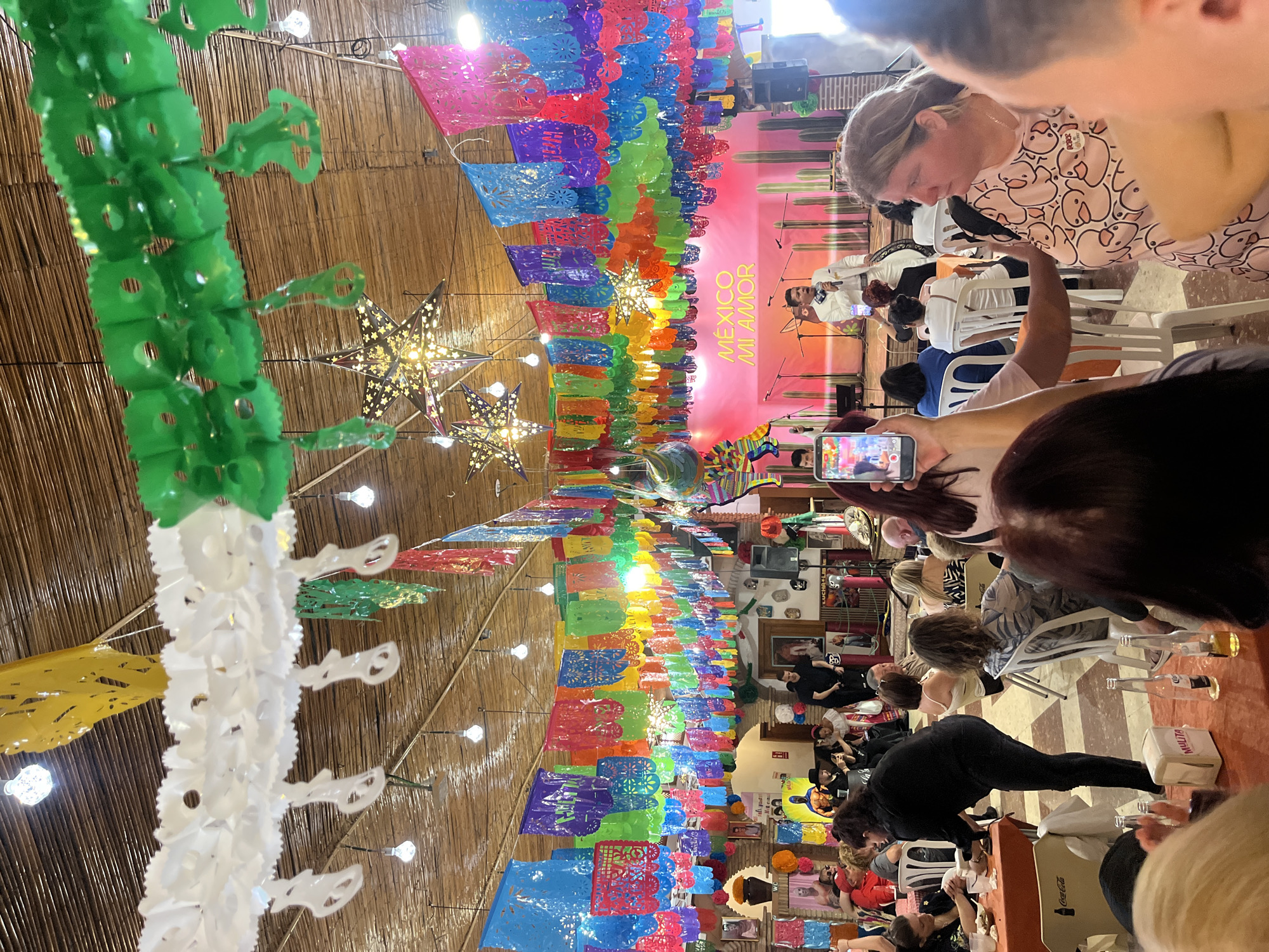 An indoor scene captures a lively Mexican-themed event in Fuengirola, Spain, adorned with vibrant decorations. Numerous strands of colorful *papel picado* (perforated paper banners) hang from the ceiling, alongside decorative green and white chains, possibly crafted from recycled plastic. Star-shaped lanterns cast a warm glow, complementing the bamboo or rattan-matted ceiling.
A crowd of people is gathered, mostly seated at tables, facing a brightly lit stage at the far end of the venue. On stage, a man in a white suit stands near microphones, suggesting a performance or presentation is underway. The stage's backdrop is a mural featuring cacti and prominently displays the text "MÉXICO MI AMOR" (Mexico My Love), reinforcing the cultural theme.
Attendees appear engaged, with several individuals holding smartphones, and at least one person actively filming the stage. In the foreground, bottles (likely beverages) and tissue boxes with "MULTIA" text are visible on tables, alongside a "Coca-Cola" branded box. The event has an evening ambiance, illuminated by artificial lighting, creating a festive and communal atmosphere.