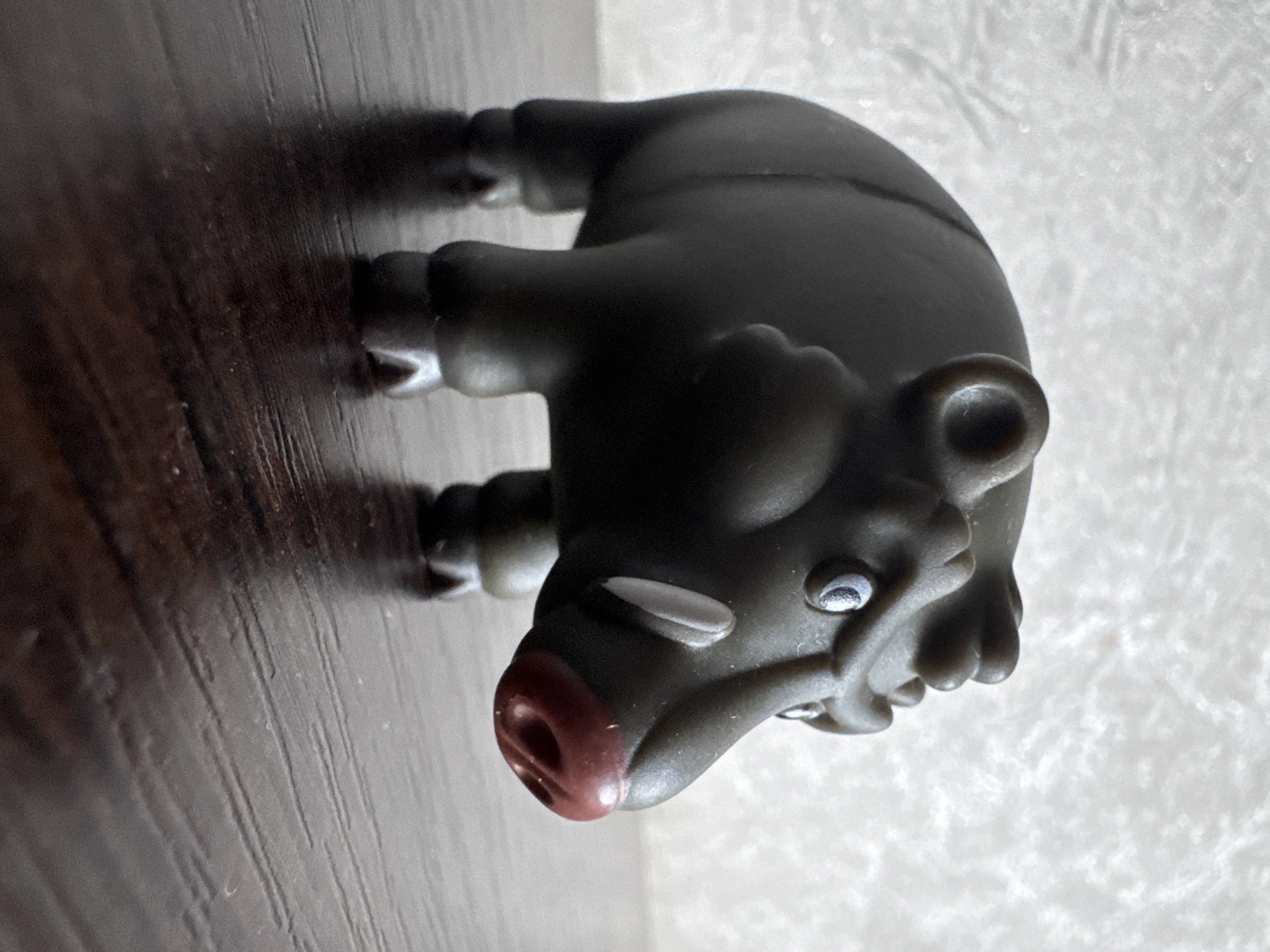 A dark gray, stylized toy boar is positioned on a dark brown wooden surface. The boar is facing to the right, with its head turned slightly towards the viewer. It has white eyes with black pupils, two visible white tusks protruding from its mouth, and a reddish-brown snout with two nostrils. The boar's body is rounded and stout, with four short legs. The background consists of a textured, light-colored surface, possibly a wall. The image is a close-up shot, with the boar occupying the majority of the frame. The lighting is soft, casting a subtle shadow on the wooden surface. The scene is indoors and appears to be a still life. There is no indication of the city of Яромінскі сельскі Савет, Belarus in this image.