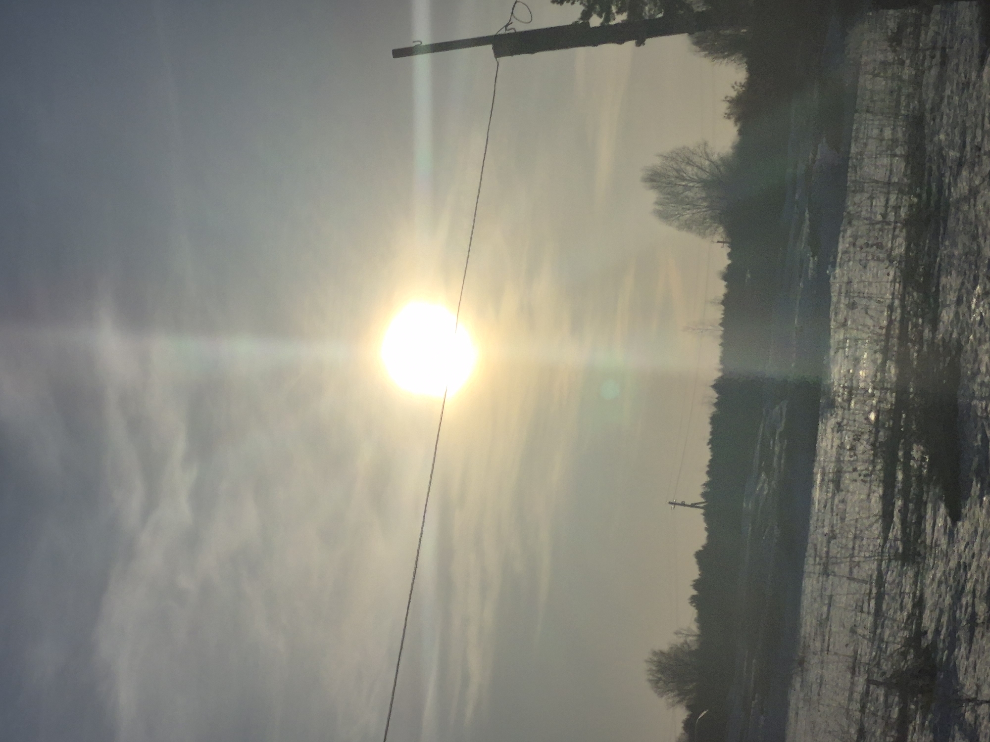 The image shows a winter scene in what appears to be the rural outskirts of a city. The sky is overcast, with the sun shining brightly through a thin layer of clouds, creating a strong lens flare. A utility pole with wires is visible in the upper left portion of the frame. The landscape is covered in snow. A line of trees is visible in the background. The location could be Дулесово, Russia.