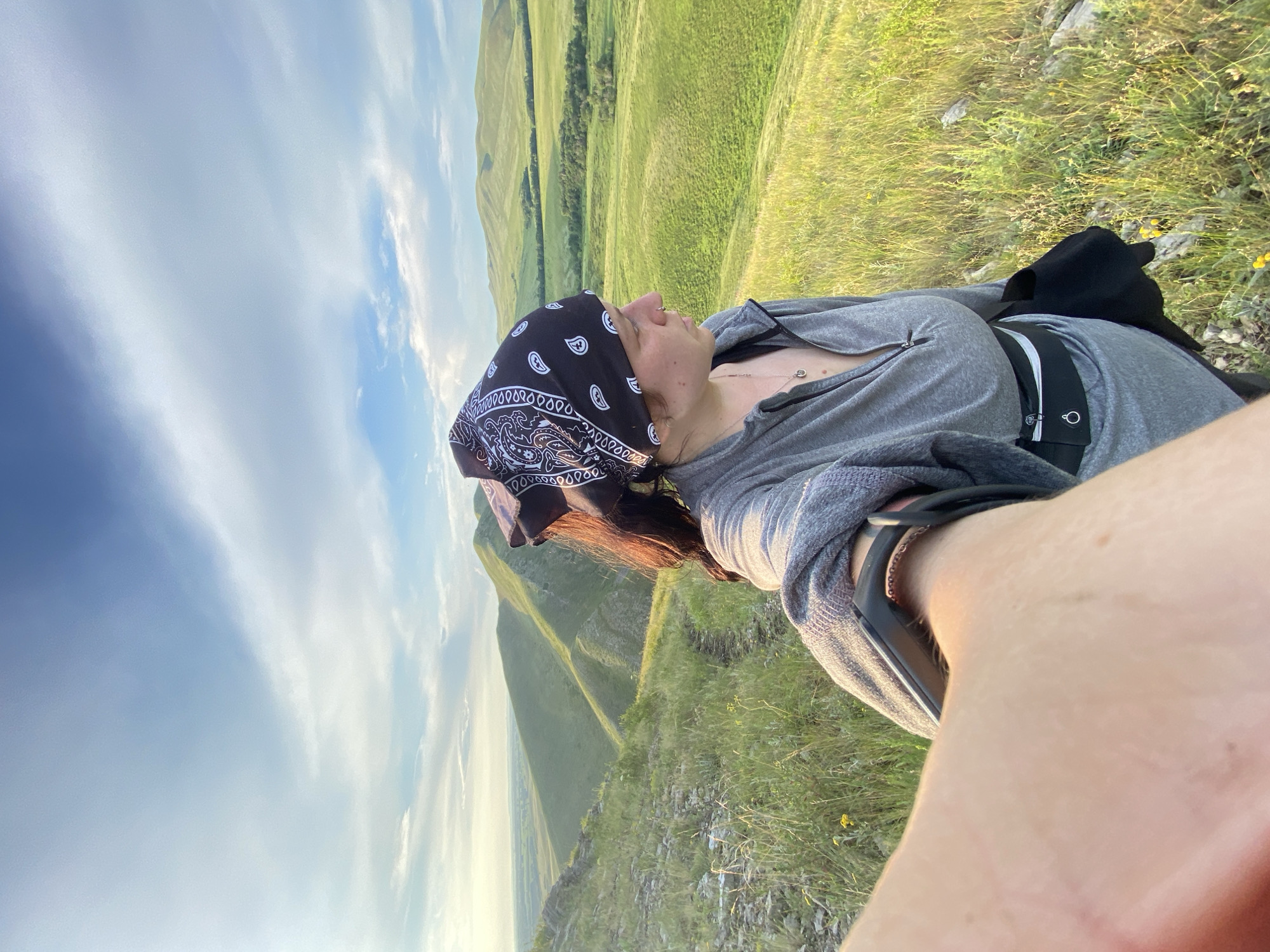 A selfie is taken by a person wearing a gray zip-up hoodie, a black bandana with a white paisley pattern, and a black fanny pack, on a grassy hillside. The person is looking to the right, with their left arm extended towards the camera, capturing a portion of their arm and hand. The background features rolling green hills and a bright blue sky with wispy white clouds, indicative of a sunny day. The location is identified as near Novogafarovo, Russia. The overall scene suggests an outdoor excursion or hike in a rural, hilly landscape during daylight hours.