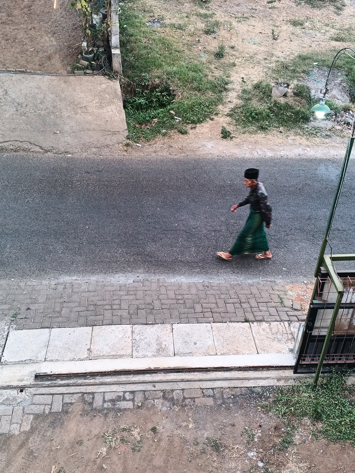 An overhead, long shot captures a man walking on a paved road in Bocek, Indonesia. The man, wearing a black peci hat, a dark long-sleeved shirt, and a green sarong, is walking towards the right side of the frame. His right leg is forward, and his left leg is behind, indicating motion. The road is an asphalt surface. To the left of the road, a concrete sidewalk with lighter rectangular concrete slabs is visible, bordered by a darker brick-like pavement. Beyond the sidewalk is a dirt area with some dry grass. To the right of the road, there is a grassy embankment with sparse vegetation and a green street lamp. A dark metal railing with a green frame runs vertically along the right edge of the image, obscuring part of the embankment. The lighting suggests it is either early morning or late evening.