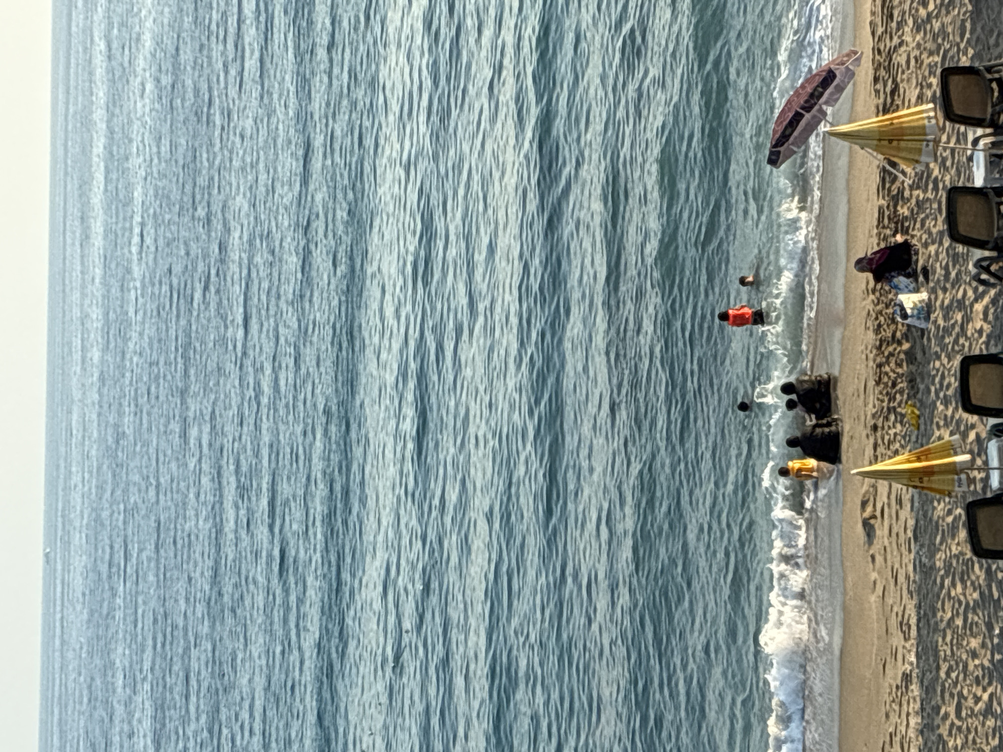 This media file shows a beach scene in Alanya, Turkey. The image is oriented vertically, with the ocean on the left and the beach on the right. Several people are visible on the beach and in the shallow water. Two people are in the water, one wearing a bright orange life vest, and another person, possibly in a yellow top, is wading near the shore. Several other figures, appearing to be sitting or lying down, are scattered on the sand.
Objects on the beach include two beach umbrellas, one a light yellow color and the other a darker, patterned umbrella. Several beach chairs are also visible, suggesting this is a developed beach area. The sand is light brown, and the water is a clear blue-green, with gentle waves lapping at the shore.
The lighting suggests it is daytime, likely late afternoon or early evening, as evidenced by the shadows cast on the sand. The weather appears to be pleasant and sunny. The overall mood of the scene is relaxed and leisurely, indicative of people enjoying a day at the beach. There are no discernible text elements within the image.