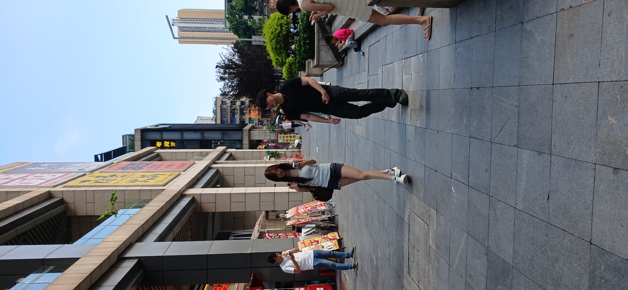 This image depicts a street scene in the Jiulongpo District of China. In the foreground, a young woman with long dark hair is walking towards the camera, looking at her phone. She is wearing a white tank top and denim shorts, with white sneakers. To her right and slightly behind her, a man with short dark hair and sunglasses is walking in the same direction, wearing a black t-shirt and dark pants. Further in the background, a man in a white t-shirt and blue jeans is standing and looking at his phone. The people are on a wide, gray tiled sidewalk in front of a modern building with a stone facade and glass railings. There are several display boards and awnings to the left of the sidewalk. In the background, other buildings and trees are visible under a clear blue sky. The scene appears to be a commercial or pedestrian area.