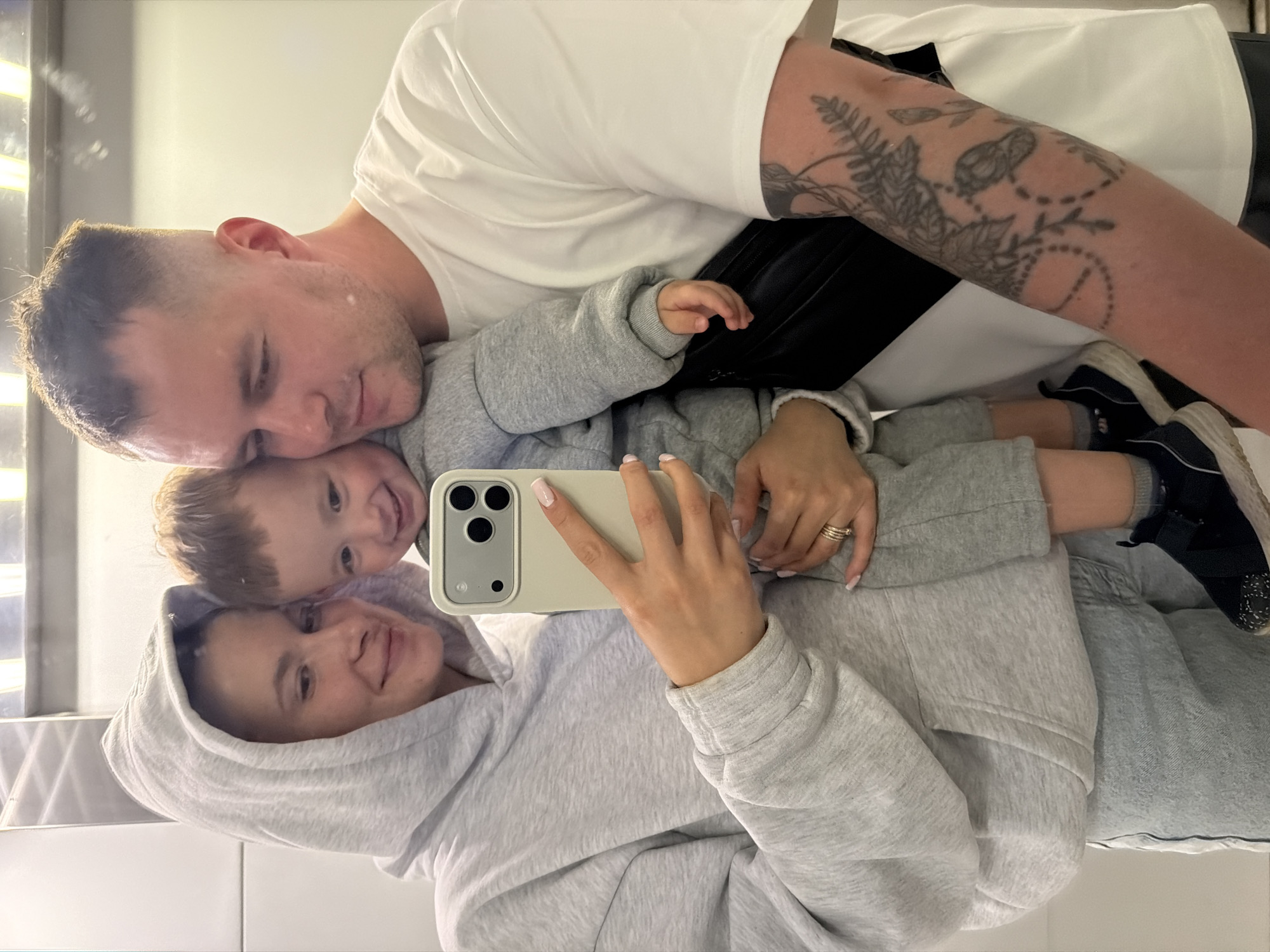 The image depicts a mirror selfie of a family of three – an adult woman, an adult man, and a toddler – taken indoors in what is described as being in the city of Odintsovo, Russia. The plain, light-colored, reflective background suggests a simple interior setting, possibly a bathroom or hallway.
On the left, an adult woman, likely in her 20s or 30s, is holding a light beige smartphone with a triple camera array in her right hand, indicating she is taking the photograph. She is wearing a light grey hooded sweatshirt with the hood up, covering her hair, and light blue denim jeans. A stack of three thin rings is visible on her left ring finger, and her nails are manicured with a light pink polish. She has a gentle smile, looking towards the phone.
In the center, a toddler, appearing to be between 1 and 3 years old, is held between the two adults. The child has short, light brown hair and is wearing a matching light grey long-sleeved top and pants, along with dark sneakers featuring white soles and grey socks. The child displays a wide, open-mouthed smile, looking directly at the phone.
On the right, an adult man, also likely in his 20s or 30s, holds the child with his right arm. He has short, dark hair with faded sides and is wearing a white short-sleeved t-shirt. A black fanny pack or crossbody bag is visible across his upper torso. His right forearm is covered with a black-and-grey tattoo sleeve featuring intricate botanical designs, including leaves, vines, beaded patterns, and what appears to be small bird or insect motifs. He is looking down at the child with a subtle, affectionate expression.
All three subjects are positioned closely, conveying a sense of family intimacy. The lighting is bright and even, illuminating the subjects clearly.
