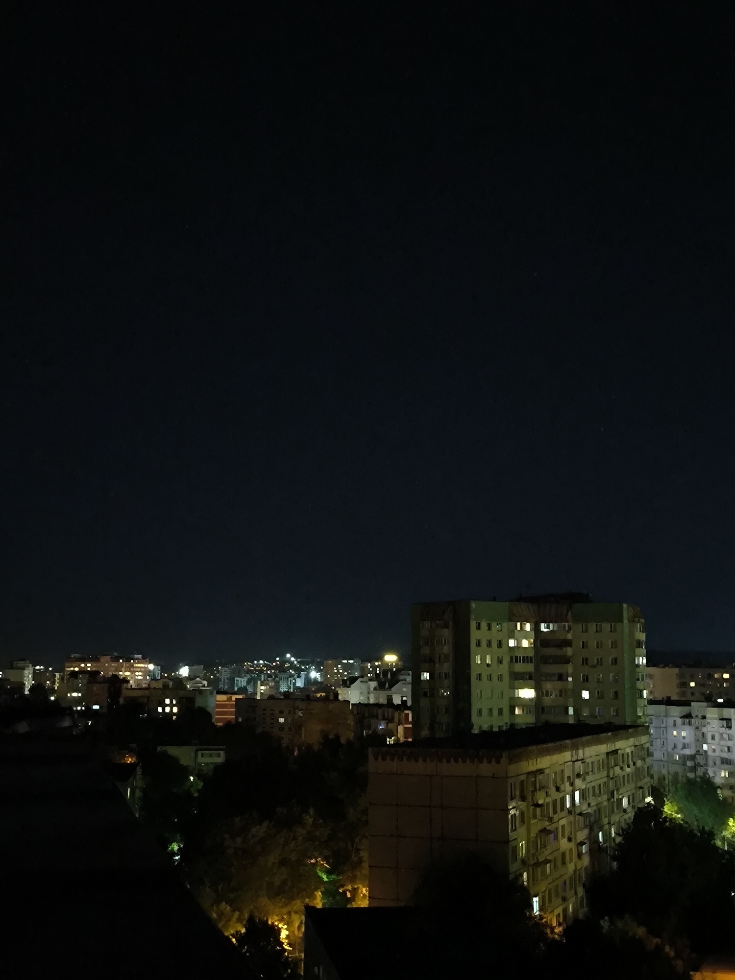 The image captures a nighttime cityscape, likely in the city of Chișinău, Moldova. In the foreground, dark rooftops are visible. The midground features several multi-story apartment buildings, with some windows illuminated, indicating occupancy. Trees are interspersed among the buildings, with some foliage illuminated by artificial light. In the background, a more densely populated area of the city is visible, with numerous buildings displaying lit windows and streetlights creating a soft glow. The sky above is dark, with a few faint stars or distant lights visible. The overall impression is one of a residential urban area at night.