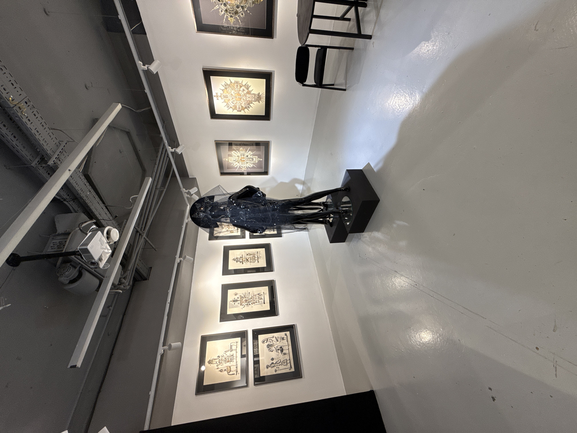 The image, originally captured in a rotated orientation, depicts an interior art gallery or exhibition space. On the right, a white wall showcases numerous framed artworks, arranged in two vertical columns. These pieces appear to be detailed drawings or prints, characterized by intricate, possibly mechanical or architectural, designs. Each artwork is set in a black frame and illuminated by track lighting fixtures mounted on the ceiling.
In the mid-ground, to the left of the art wall, a black mannequin or sculpture stands on a dark, rectangular pedestal. It is adorned with a sheer, dark garment that seems to incorporate small, reflective elements like glitter or sequins. Further to the right, a small black table and two simple, modern black chairs are visible against the white wall and floor, suggesting a small seating area.
The ceiling is industrial in design, featuring exposed grey ductwork, metal cable trays housing electrical wires, and various conduits. Multiple white track lights and a projector-like device are mounted on the ceiling, contributing to the overall artificial illumination of the space. The floor is light-colored, possibly white or a very pale grey, with a glossy finish that reflects light and shows some minor scuffs. The environment is clean and minimalist, designed to draw attention to the displayed art. There are no people visible, and no discernible text is present in the image.