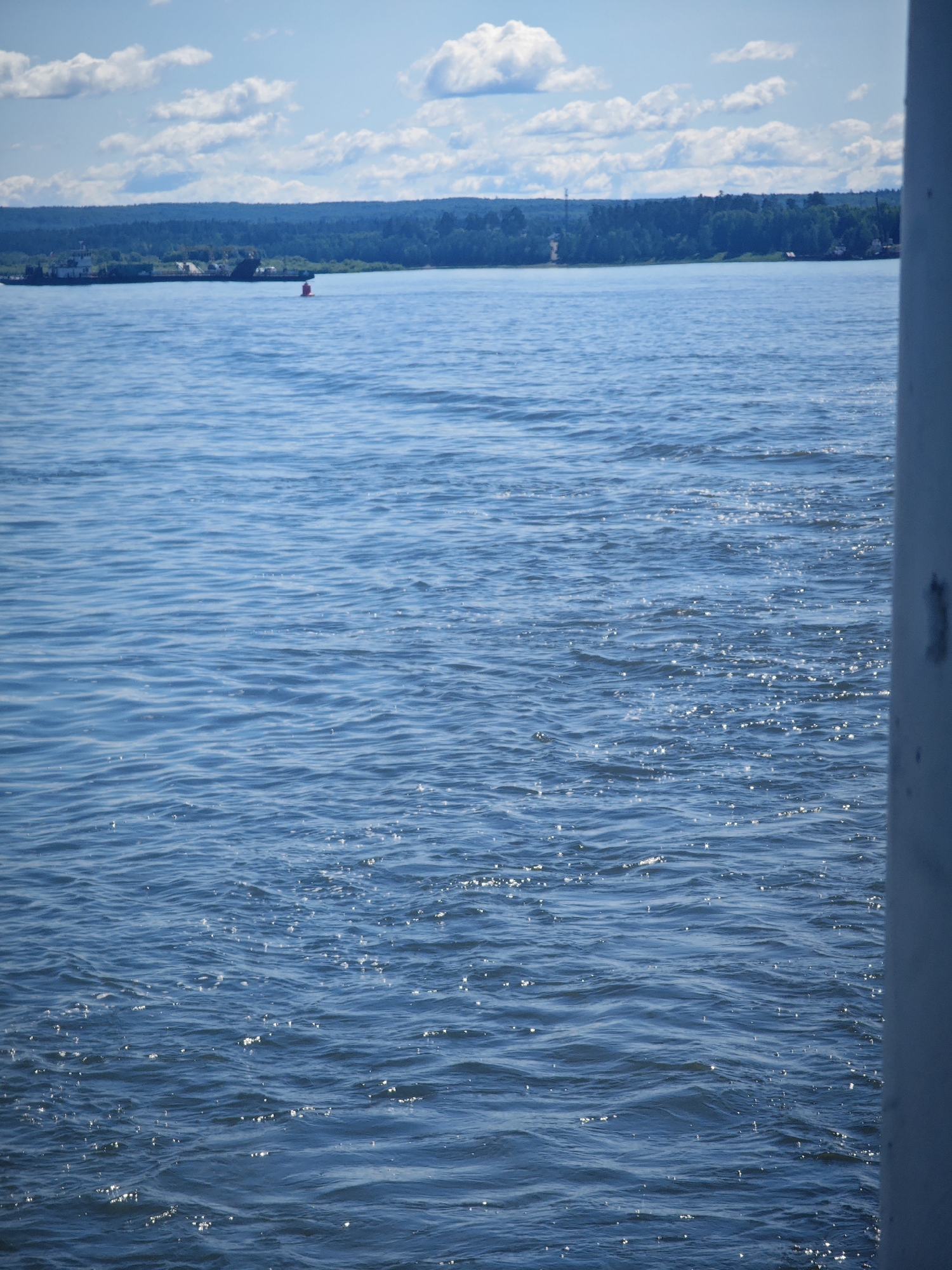 The image shows a body of water with a distant shoreline lined with trees and some buildings. The sky is blue with scattered white clouds. The water is rippled and reflects the sunlight, creating sparkling highlights. A red buoy is visible in the water, as well as a barge or boat further in the distance near the shore. The right side of the image is partially obstructed by a vertical, light-colored object, possibly a railing or wall of a vessel. The scene is serene and natural, evoking a sense of a peaceful day on the water. The provided context suggests the location is Krutoy Log, Russia.