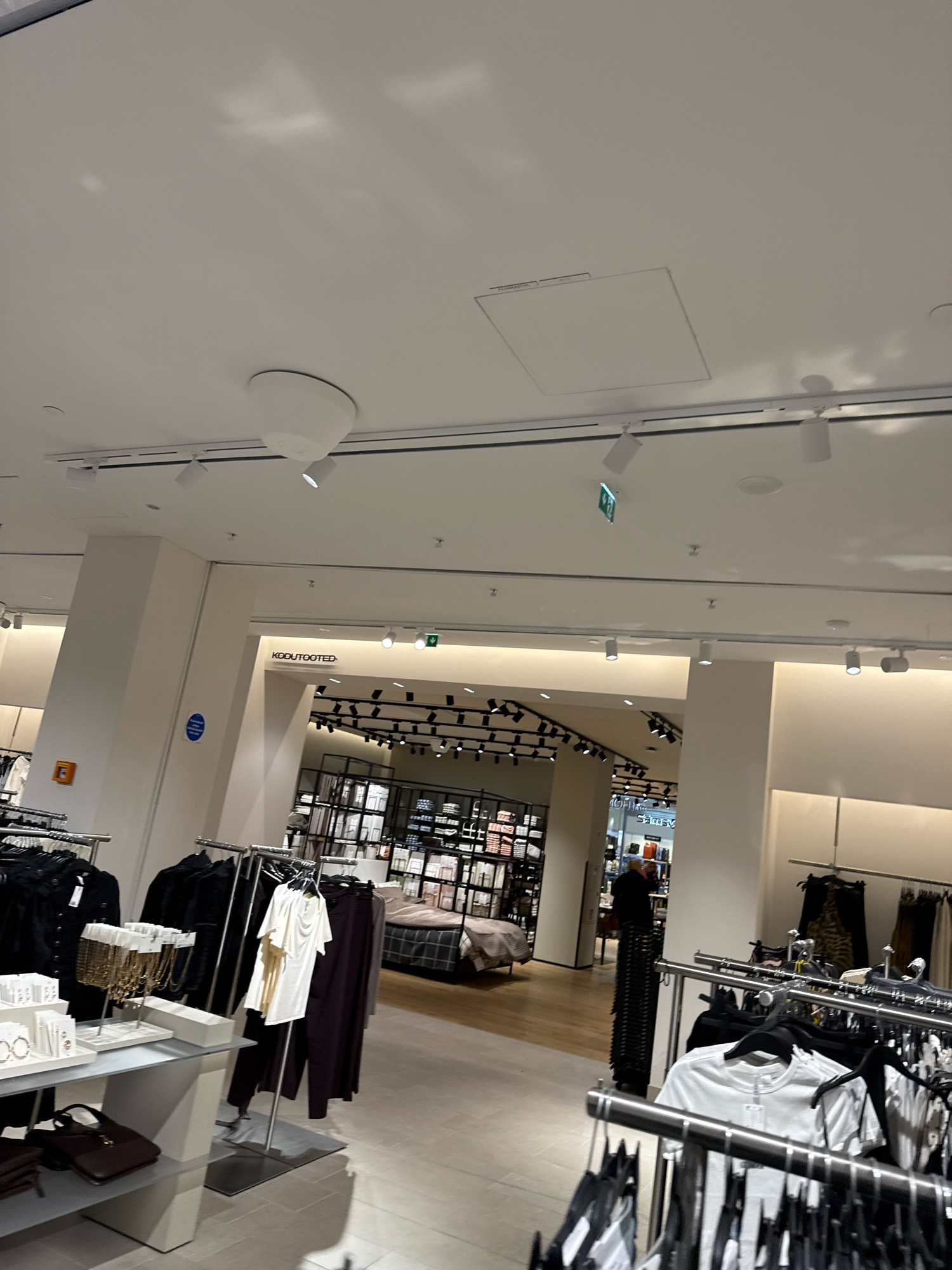 The image depicts the interior of a modern retail store, likely a clothing boutique or department store, located in Tallinn, Estonia. The store features a bright, well-lit environment with white ceilings and walls, and displays of clothing on racks and shelves. Prominently visible are rows of garments, accessories, and mannequins. In the background, a sign reads "KODUTOOTED," which translates to "Home Products" in Estonian, suggesting a section of the store dedicated to home goods or perhaps the store's name.
The ceiling is a key feature, showing a recessed panel, track lighting, and a large, circular diffuser for ventilation or lighting. One spotlight is angled downwards, and a green emergency exit sign is visible. The flooring appears to be a light-colored tile or laminate.
In the foreground, clothing racks are filled with garments. On the left, a display table showcases accessories like necklaces and earrings, and some handbags are visible on a lower shelf. Further back, the store opens up to reveal more clothing displays and what appears to be a bed display, suggesting a home furnishing section.
A few individuals are present in the store. One person, dressed in dark clothing, is standing near the entrance to the "KODUTOOTED" section, looking towards the interior. Another person is partially visible in the background, near more retail displays. The overall impression is of a clean, organized, and contemporary retail space.