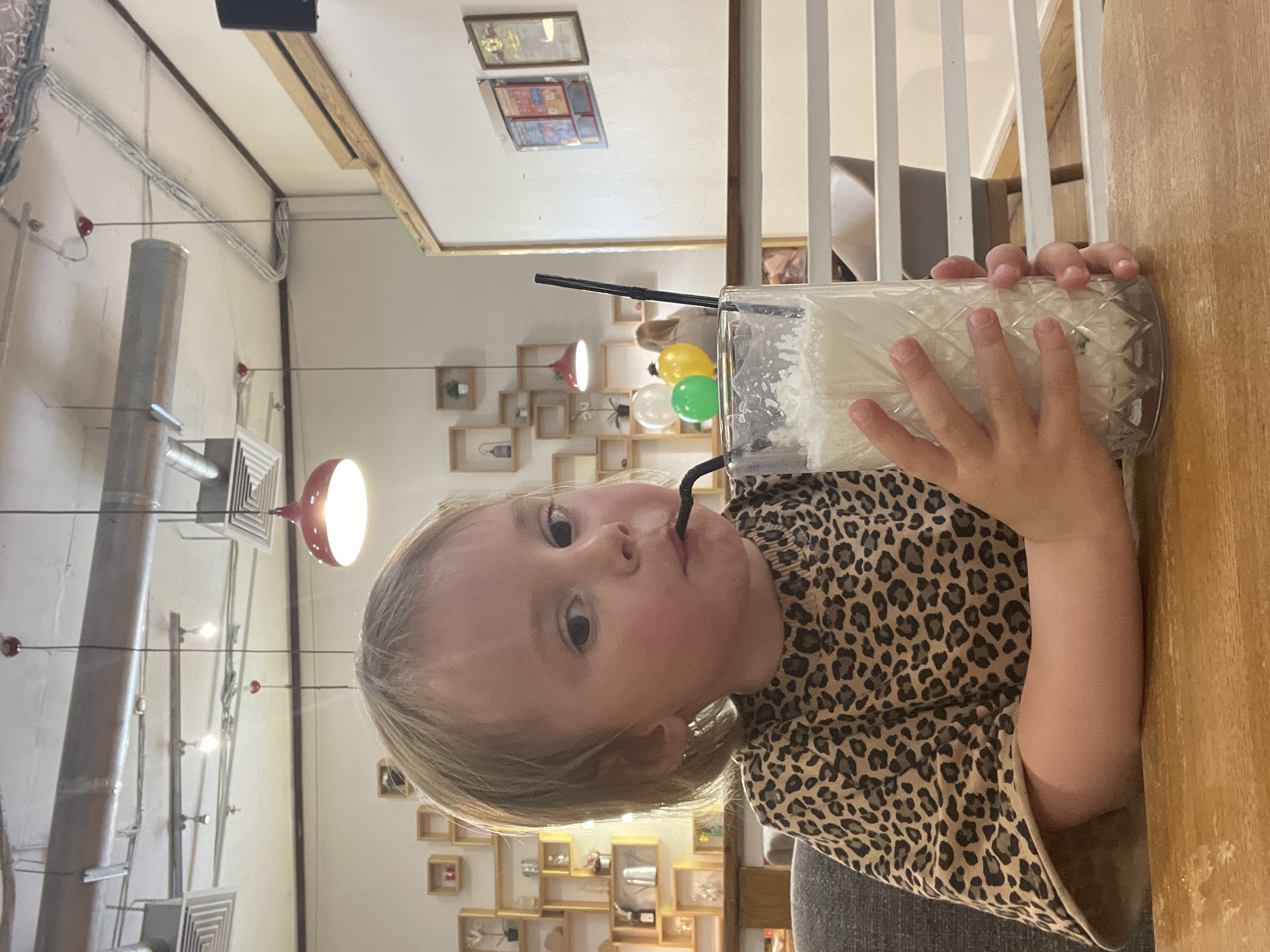 A young Caucasian child with blonde hair is sitting at a wooden table, drinking a milkshake from a tall glass with a black straw. The child is wearing a brown leopard-print shirt. The background shows a cafe interior with shelves on the wall, red pendant lights, and a whiteboard with framed pictures. There are also balloons in the background. The overall setting suggests a casual indoor dining establishment. The image is taken from a medium close-up perspective, focusing on the child and the drink.