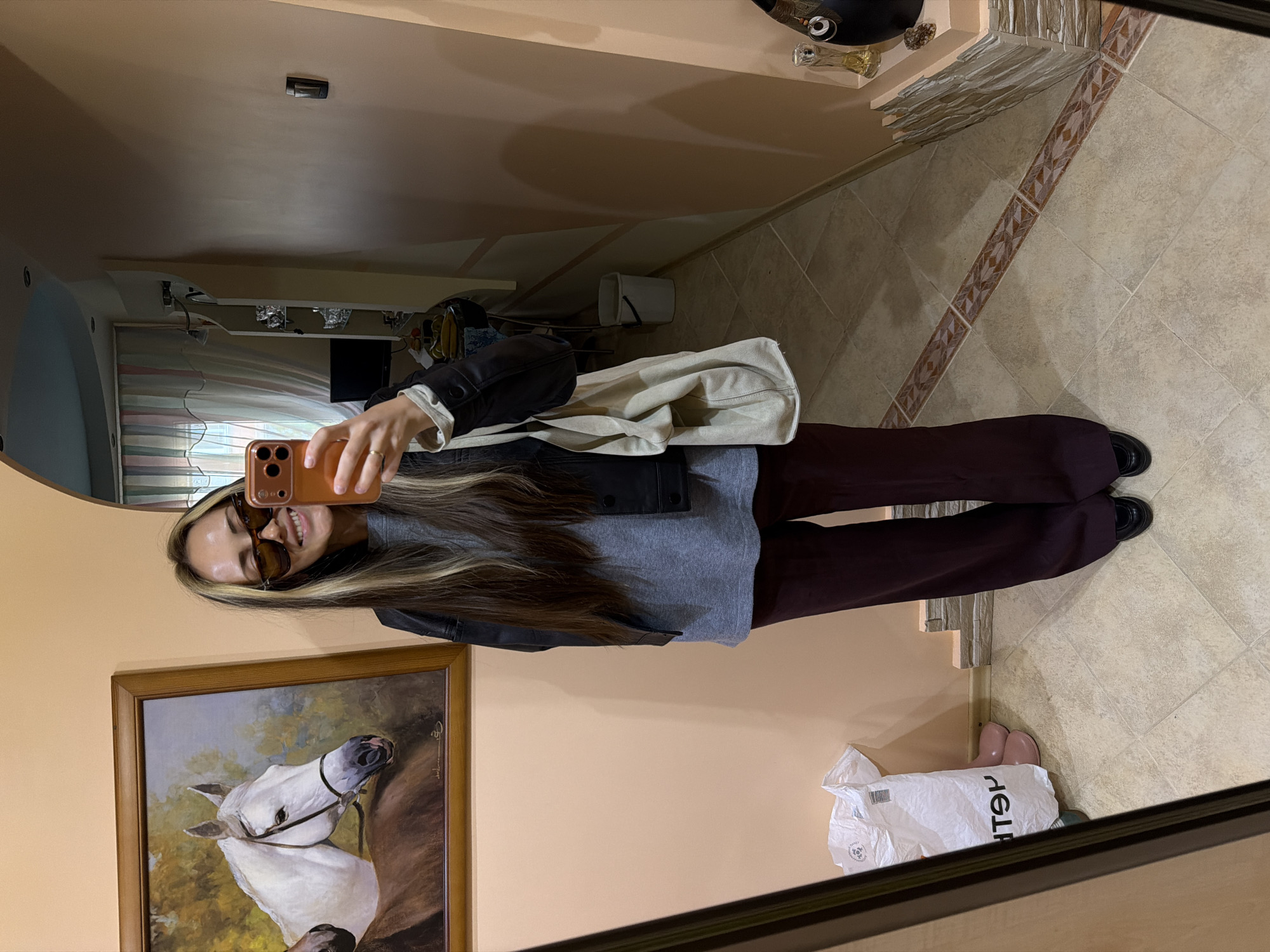 The image shows a young woman taking a selfie in a mirror. She is wearing sunglasses, a grey top, a black jacket, and dark maroon pants. She is holding an orange phone. A large painting of a white horse in a brown frame is visible on the wall behind her. A white shopping bag with black writing is on the floor in the lower right corner of the image. The floor is tiled in light beige and has decorative brown and beige tiles. There is no discernible evidence in the image to confirm the location in Kamyshin, Russia.