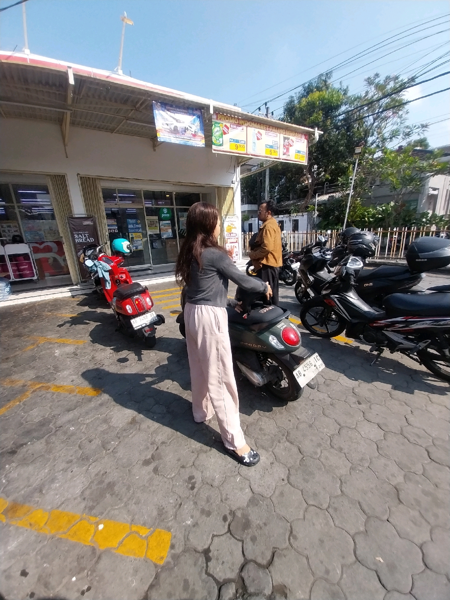 The image depicts a scene in front of a convenience store in Depok, Indonesia. A woman with long brown hair, wearing a grey long-sleeved shirt and light pink pants, is standing in the foreground, facing away from the camera and towards a red scooter. She is wearing black and white flats. A man wearing a tan shirt and black pants stands to the left, further back, near the entrance of the store. Several other motorcycles are parked in the lot. The store has a white facade with a red awning and displays promotional banners. The ground is paved with interlocking concrete blocks, and yellow lines mark parking spaces. The sky is clear and blue, indicating a sunny day.