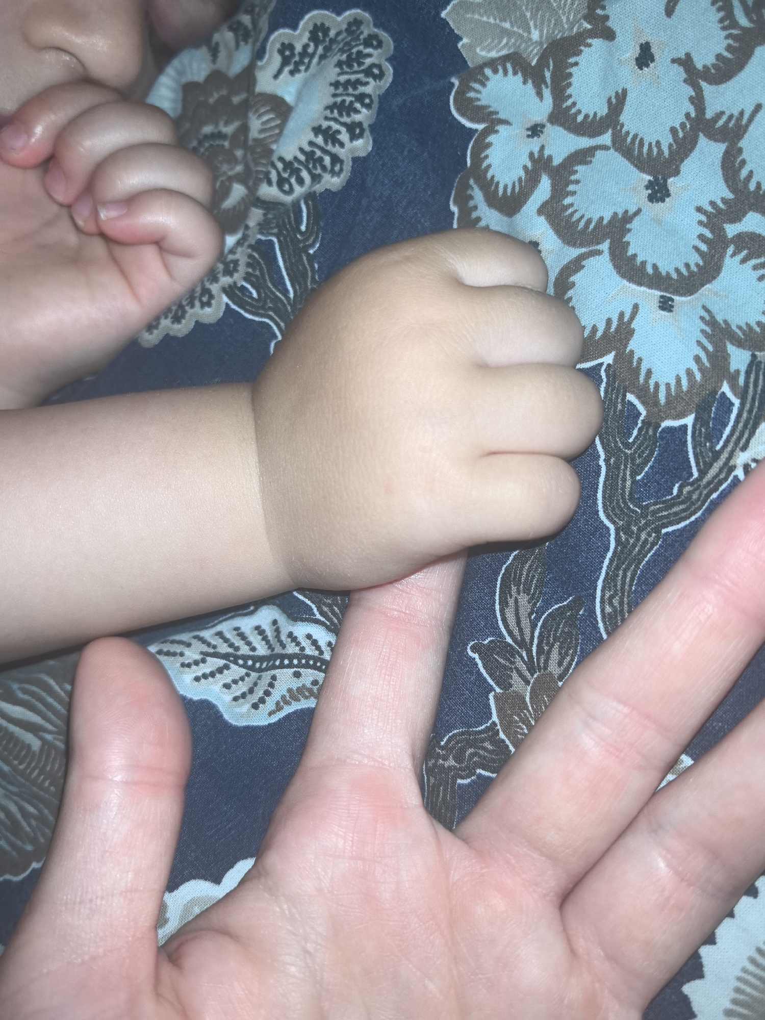 A close-up photograph shows two hands, one significantly smaller than the other, placed on a surface with a floral pattern. The smaller hand, presumably a baby's, is resting on the index finger of the larger hand. The location is Unknown, Unknown.
