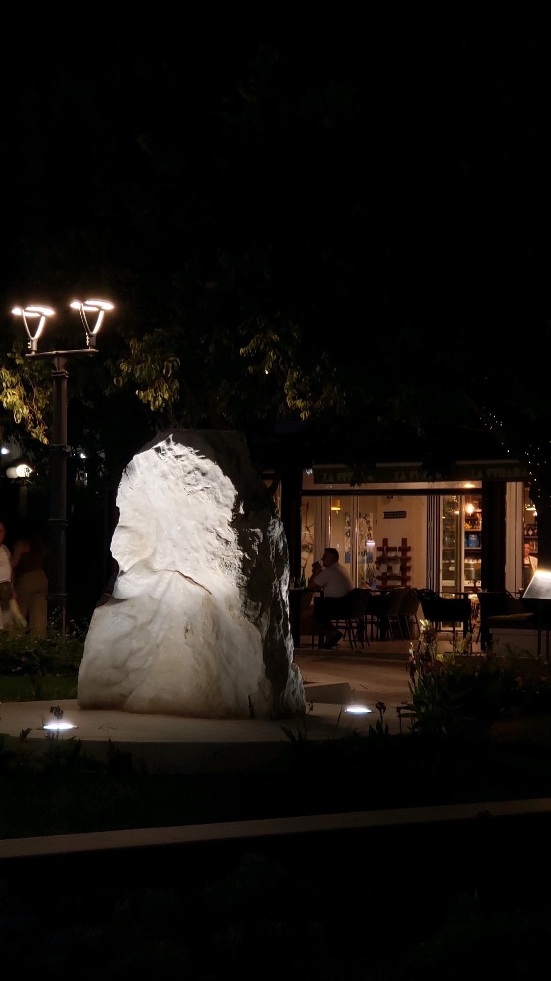 This is a night-time, eye-level, full-frame shot taken in Općina Tučepi, Croatia, depicting an outdoor seating area of a restaurant. In the foreground, a large, illuminated white rock sculpture is the dominant feature. Behind the sculpture, a restaurant with glass doors and windows is visible, with patrons seated at tables. Two streetlights are also present, illuminating the scene. The environment suggests a relaxed evening atmosphere in a tourist area.