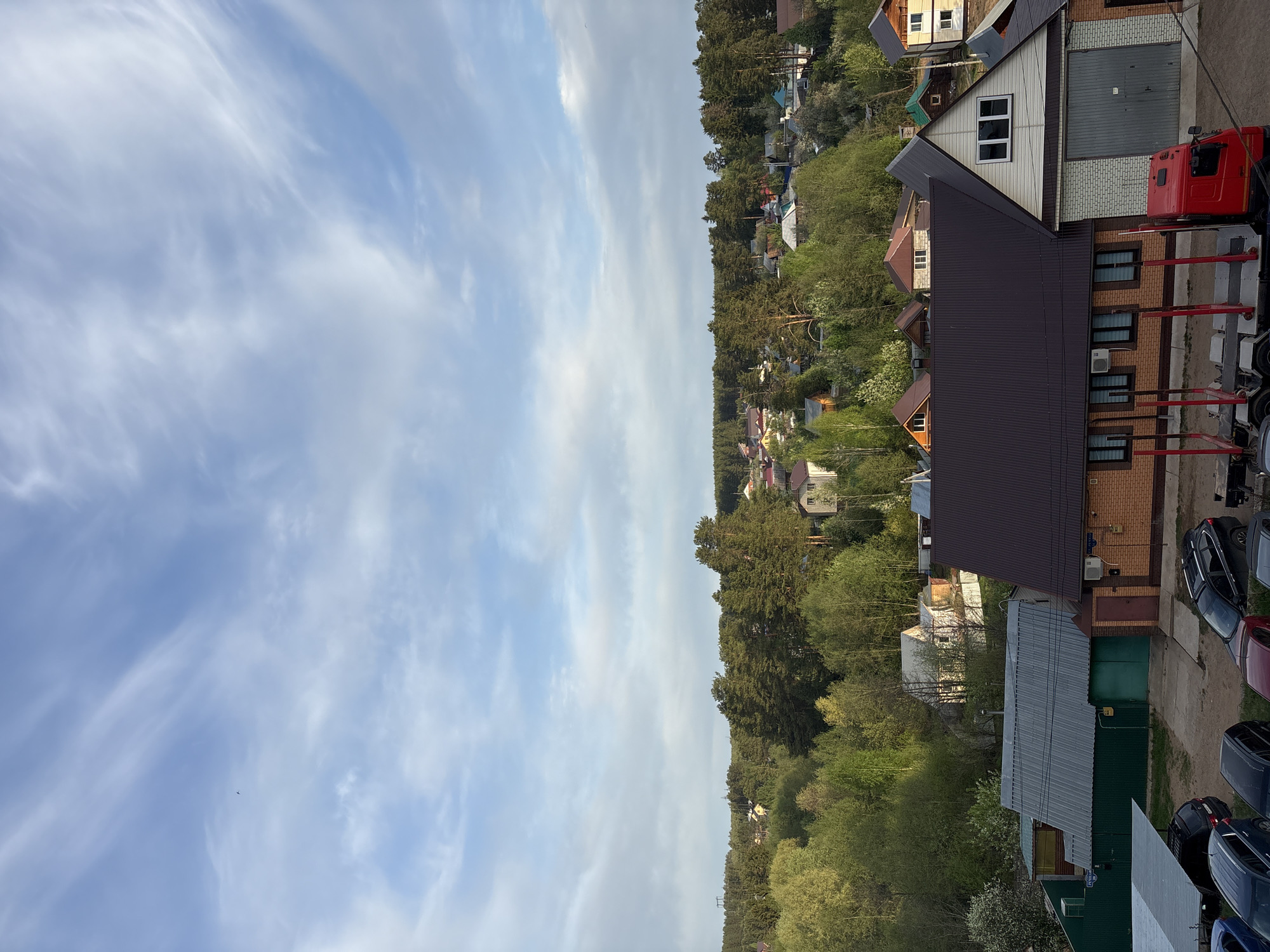 The image shows a view over a residential area, possibly in Chaikovsky, Russia. The scene is dominated by the sky, with light blue tones and wispy clouds. Below, a dense band of trees and foliage separates the sky from the residential buildings. The architecture is mixed, with houses of varying styles and materials, including brick, wood, and metal roofing. Some buildings have garages and are situated on what appear to be private lots. Several cars are parked along a paved area next to some buildings, indicating residential or commercial activity. A red truck is partially visible. The lighting is soft, suggesting either early morning or late afternoon. No people are visible. The environment is a mix of natural and built elements.