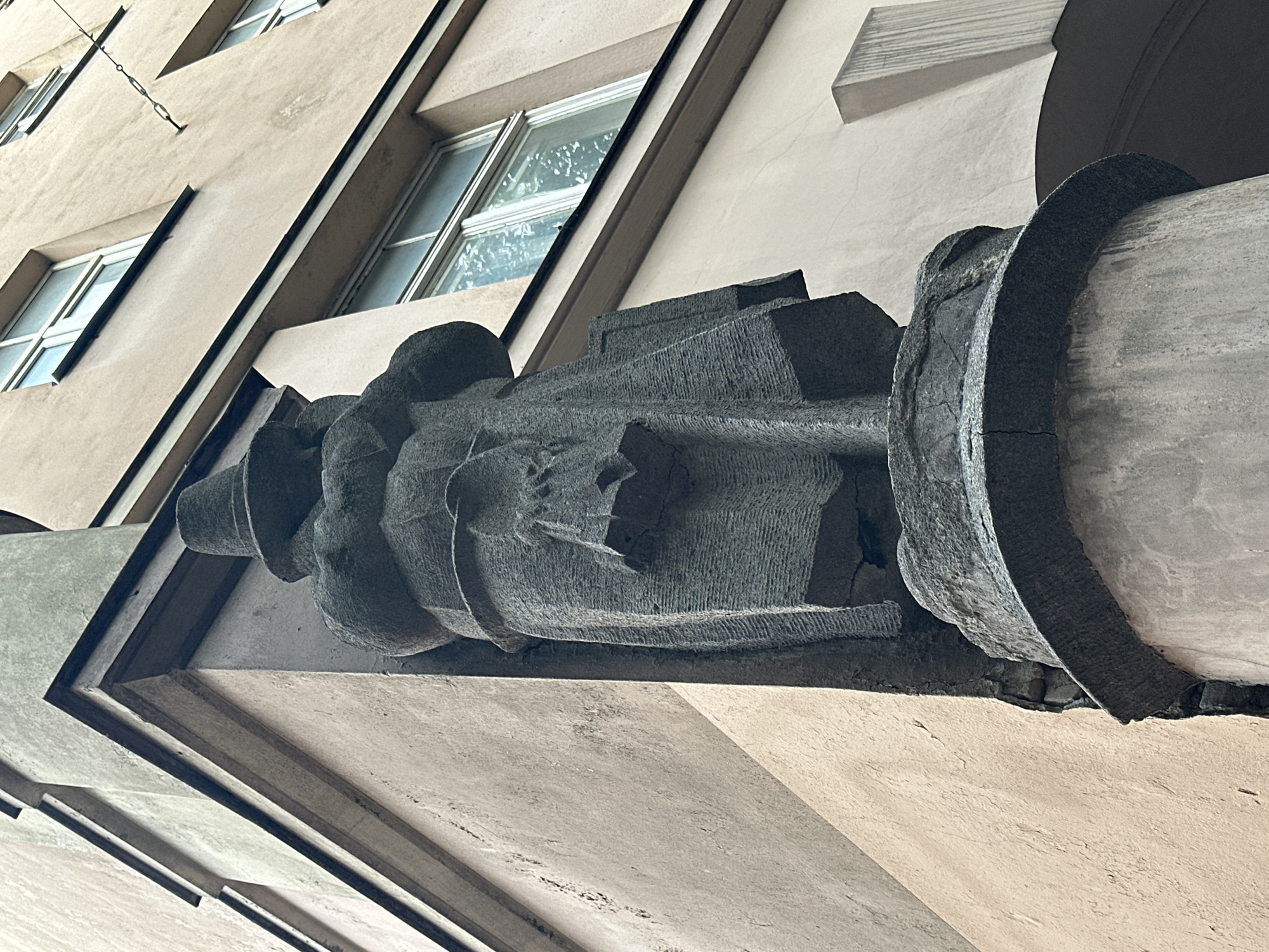 This is a low-angle, medium shot of a stone sculpture of a person wearing a hat and a long coat, positioned on the corner of a building. The sculpture is dark gray and appears to be weathered, with visible texture and cracks. The building is light beige with multiple windows, some of which reflect the sky. The architectural style suggests an older European city. The sculpture is located on the corner of Jauniela Street in the Old Town of Riga, Latvia, as part of the historic architecture.