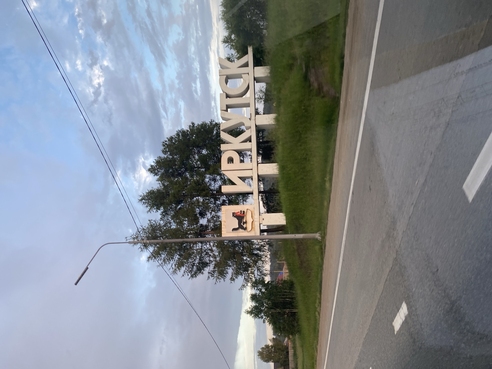 A welcome sign to Irkutsk, Russia, stands on the side of a road. The sign consists of large, white, three-dimensional letters spelling out "ИРКУТСК" (Irkutsk). To the left of the letters is a crest with a black sable holding a red bow. The sign is situated on a grassy hill, with trees behind it. A lamppost stands to the left of the sign. The road in front of the sign has white lane markings. The sky is partly cloudy. The overall scene suggests the outskirts of a city or town.