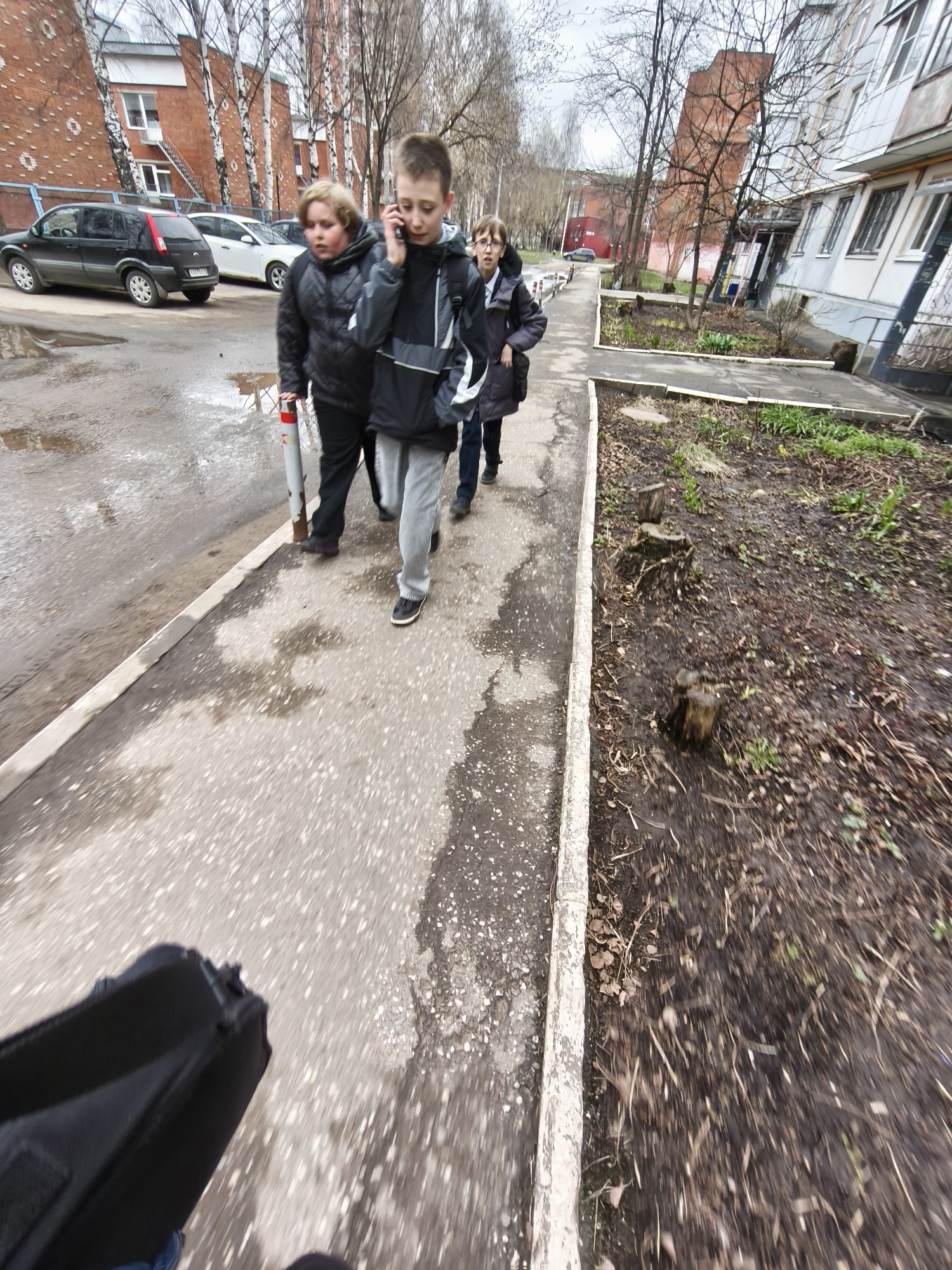 The image captures three young boys or teenagers walking along a concrete sidewalk in what appears to be a residential area in Izhevsk, Russia. The boy in the foreground, on the right, wears a grey and black jacket and is actively engaged in a phone call, holding a smartphone to his ear. To his immediate left, another boy with a fuller face is dressed in a black puffer jacket and dark pants, holding a red and white striped pole or bollard. A third boy, wearing glasses and a dark jacket, is partially visible behind the second boy.
The primary activity is the boys walking together. The scene suggests a typical, everyday occurrence.
Notable details include the overcast weather, indicated by the lack of strong shadows and diffused light. The ground next to the sidewalk is muddy and unkempt, with sparse patches of grass, leaf litter, and several visible tree stumps, suggesting either early spring or late autumn/winter. Puddles are present on the asphalt road next to the sidewalk, further indicating damp conditions. Bare deciduous trees line the background, and multi-story apartment buildings, one with red brick and another with a lighter facade, frame the street. The perspective is low, possibly from someone walking behind and slightly to the side of the group, with a black bag visible in the bottom left corner of the frame. There is no clearly legible text on any objects or buildings within the image. The overall atmosphere is neutral, reflecting a common urban environment on a drab day.