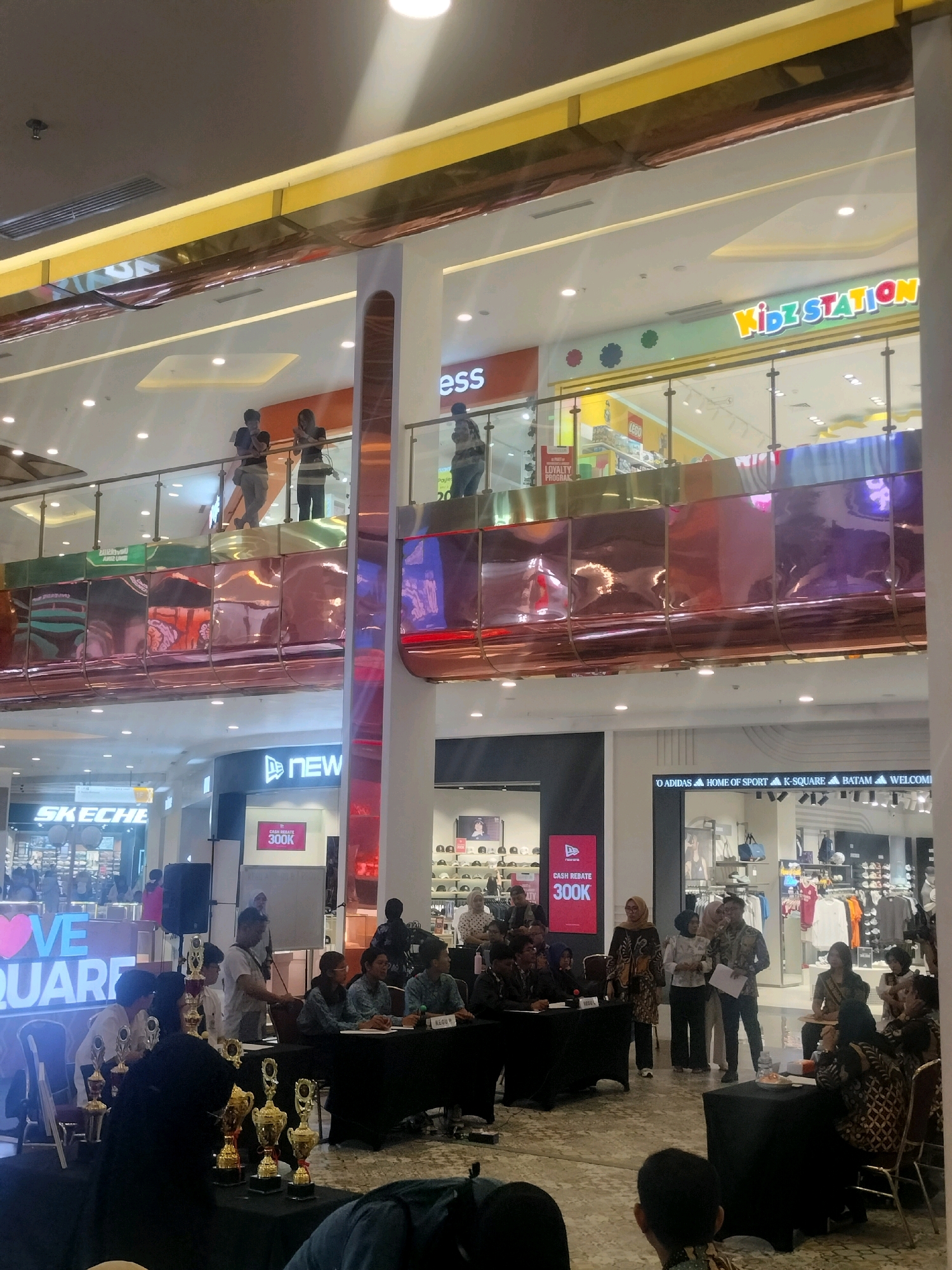 The image depicts an indoor scene, likely a shopping mall in Batam, Indonesia, during an event or competition. In the foreground, several people are seated behind a long black table covered with trophies, suggesting an awards ceremony or judging panel. The individuals appear to be participating in a formal event, with some dressed in traditional batik attire.
Behind the table, a group of participants and organizers are standing, some holding papers and appearing to be in discussion or presenting information. The environment suggests a public space with various retail stores visible in the background. Signs for "SKECHERS" and "NEW ERA" are clearly visible on the storefronts, indicating a commercial setting.
Further in the background, on an upper level of the mall, another group of people are standing near a railing. A brightly lit sign for "Kidz Station" is prominent, suggesting the presence of children's retail or entertainment venues within the mall. The mall's interior features modern architecture with reflective surfaces and warm lighting, creating a spacious and well-lit ambiance. The overall scene suggests a lively event taking place in a modern shopping complex.