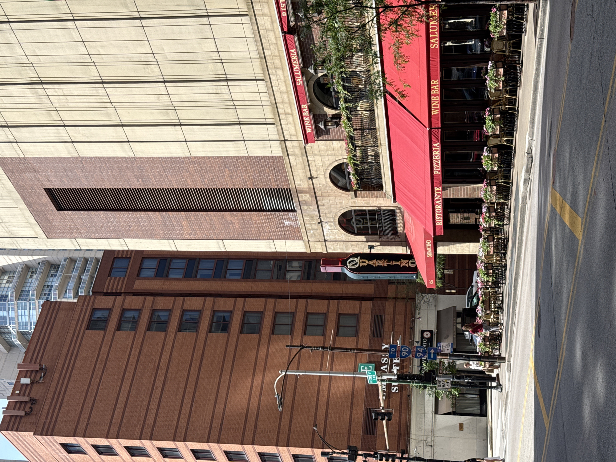 This is an image taken in an urban setting, possibly Chicago, USA, showing a street scene with several buildings and a red awning extending from one of them. The perspective suggests the photo was taken from a slight downward angle.
The buildings vary in architectural style. On the left, a modern building with a light-colored facade features a grid-like pattern. Adjacent to it is a brick building with a dark, rectangular vent. Further to the right, another brick building, this one with darker red bricks and vertical grooving, has several windows. In the background, a building with a glass facade is visible.
A prominent red awning covers the outdoor seating area of a restaurant. The awning has text indicating "RISTORANTE PIZZERIA WINE BAR SALUMERIA" and the restaurant name "QUARTINO" printed on it in gold lettering. Outdoor tables and chairs are set up along the sidewalk, decorated with potted plants. A street sign is visible, with directional arrows indicating "TO 90" and "94", suggesting a highway nearby. Another sign reads "ONE WAY".
The time of day appears to be daytime, with bright sunlight casting shadows. The weather seems pleasant, with trees and plants suggesting a warm season. A few people are visible, one walking on the sidewalk and others potentially seated at the restaurant. The overall scene portrays a lively urban street with a dining establishment and its outdoor seating.
Visible text includes restaurant names and descriptions like "QUARTINO", "RISTORANTE", "PIZZERIA", "WINE BAR", "SALUMERIA", highway designations "90" and "94", and "ONE WAY".