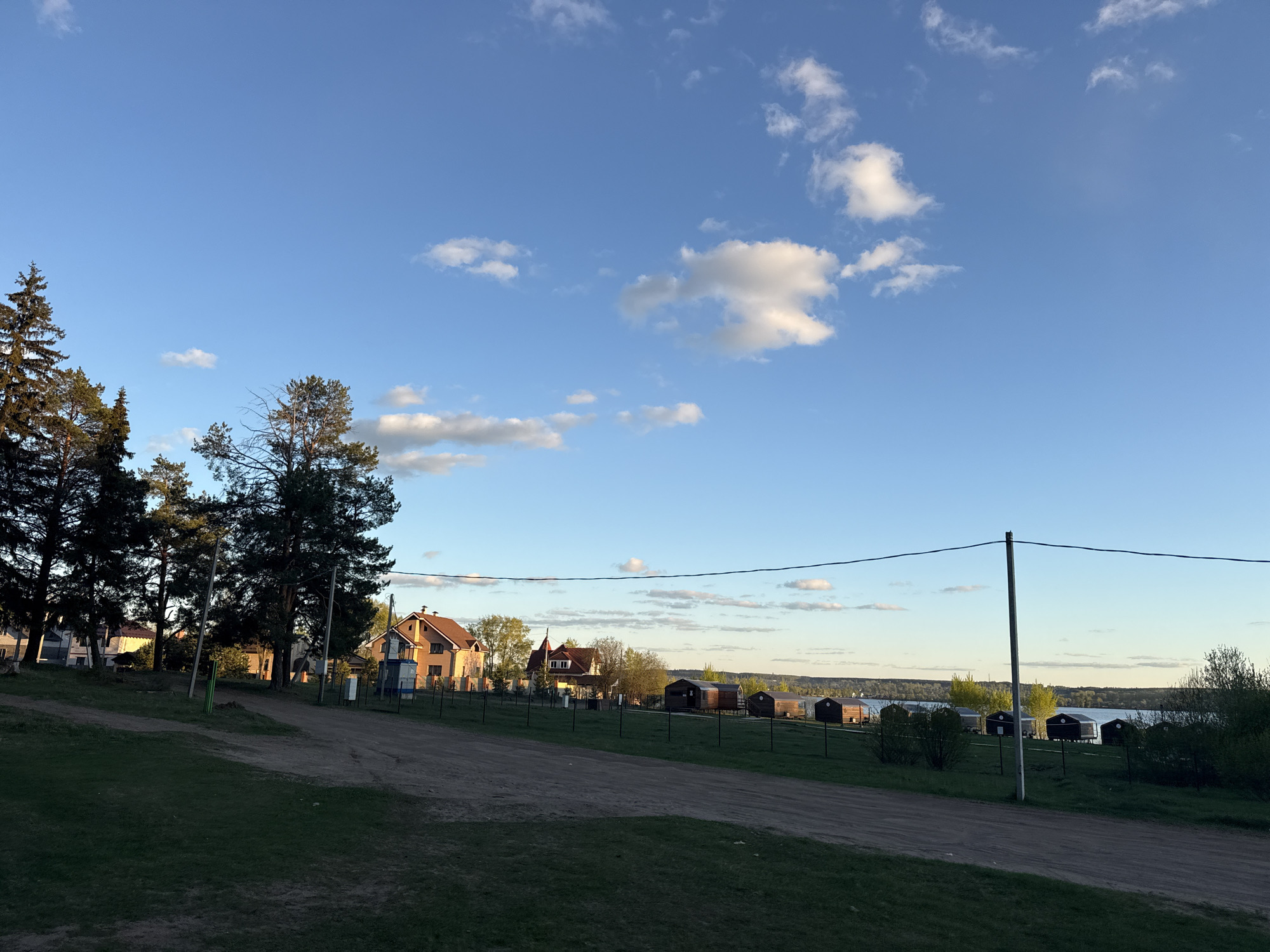 The image shows a rural scene in Votkinsk, Russia. A dirt road runs through a grassy area, bordered by trees on the left. Beyond the trees and across the road are houses and small wooden cabins on stilts near a body of water, likely a lake or river. A fence runs along the edge of the properties. Power lines stretch across the scene. The sky is blue with scattered white clouds. The lighting suggests it's either early morning or late afternoon. There are no people visible in the image.