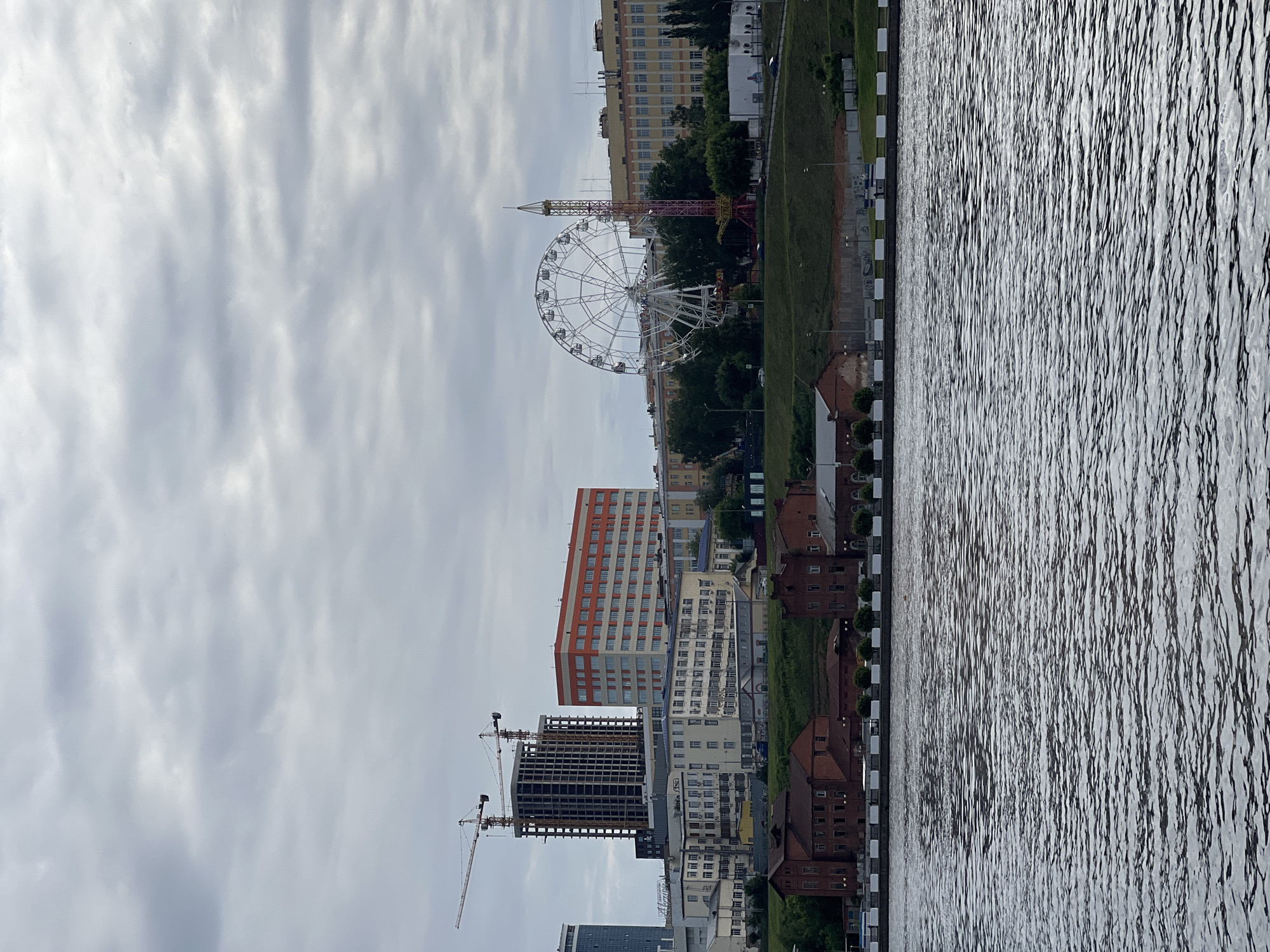 The image presents a panoramic view of the city of Izhevsk, Russia, seen from across a body of water, likely the Izhevsk Pond or a river flowing into it. The scene unfolds under an overcast sky filled with diffuse, gray cloud cover.
In the foreground, the water surface exhibits numerous small ripples, indicating a gentle current or breeze, reflecting the subdued light of the sky. Directly across the water, a paved embankment is visible, featuring a low barrier composed of alternating white and dark gray/black rectangular blocks.
Above the embankment, a strip of green, grassy slope ascends towards the city proper. At the base of this slope, several red-brick buildings with multiple windows are situated, appearing to be older structures. Further up the slope, scattered trees and shrubs are present.
The cityscape itself, stretching from the mid-ground to the background, showcases a mix of architectural styles and urban development. On the left side, a tall, multi-story building is under construction, its concrete frame prominently visible, flanked by two large tower cranes. Adjacent to it, another modern, lighter-colored high-rise building stands.
Towards the center-left, several multi-story buildings with varied facades are visible; one distinct building features horizontal orange and white stripes. These are interspersed with older, lighter-colored structures, likely residential or administrative.
The center-right of the cityscape is dominated by leisure attractions: a large white Ferris wheel is partially obscured by trees, and directly beside it stands a tall, slender amusement ride with a colorful (red, yellow, and pink) spire-like structure, consistent with a drop tower. Beyond these, more multi-story buildings, predominantly light yellow or beige, complete the urban skyline. Roads with a few vehicles are visible snaking through the developed areas.
The overall atmosphere is calm and subdued, characteristic of an overcast day. No individuals are clearly discernible due to the distance.