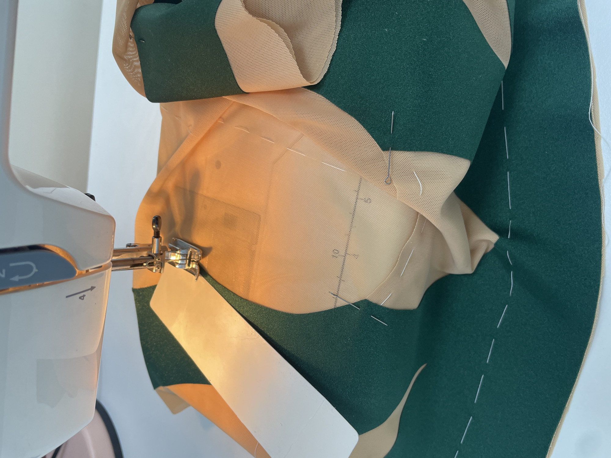 A sewing machine needle is shown poised above a piece of tan mesh fabric and green fabric, held together by pins and temporary white stitching. A white paper guide is positioned under the needle. The scene is lit by an overhead light source that casts warm tones on the fabric. The background is out of focus but suggests a clean, indoor workspace. The specific location of Pargolovo, Russia is not evident from the visual information provided in the image.