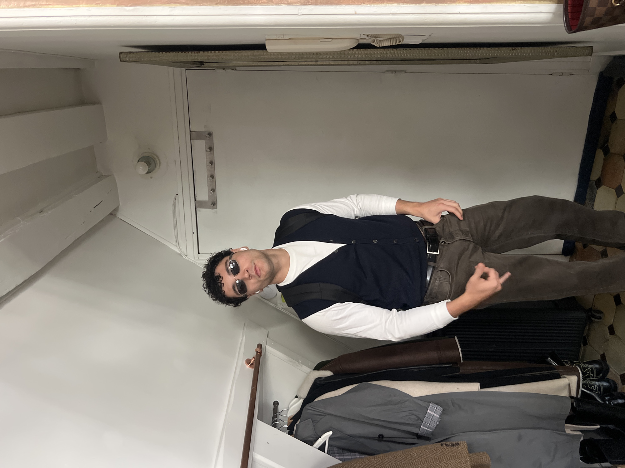 A young man with curly dark hair and sunglasses stands in a narrow hallway. He is wearing a white long-sleeved shirt, a dark blue vest, and brown pants. He has a backpack on and is holding a cell phone. To his left, there is a coat rack with several jackets hanging on it, and a dark suitcase. To his right, there is a white door with a handle and a beige intercom. The hallway has a white ceiling with exposed beams and a tiled floor. The image appears to have been taken in Paris, France.