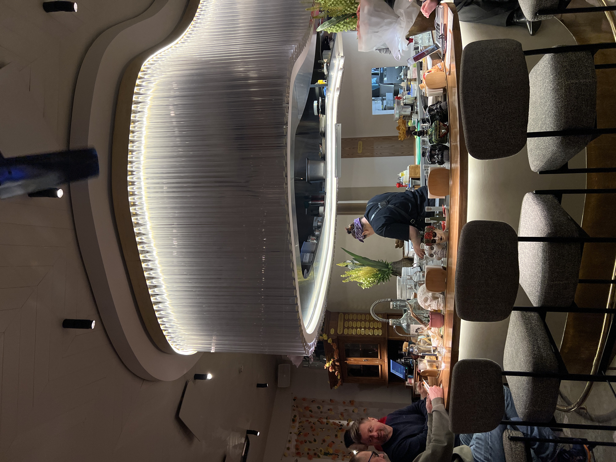 The image depicts the interior of a restaurant or bar, likely located in Moscow, Russia. A prominent feature is a large, ornate circular chandelier made of numerous clear glass or plastic rods hanging from a rounded frame with light strips. Below the chandelier is a curved bar with bar stools. A person wearing a black uniform is behind the bar, possibly preparing drinks or food. On the bar are various items, including flowers, bottles, glassware, and decorative objects, some with a skull motif. Two people are seated at the bar, one of whom appears to be looking at a phone or tablet. There are decorative elements on the walls, including wallpaper, lights, and an antique-looking cabinet. The bar stools are upholstered in a gray fabric and have a black metal frame.
