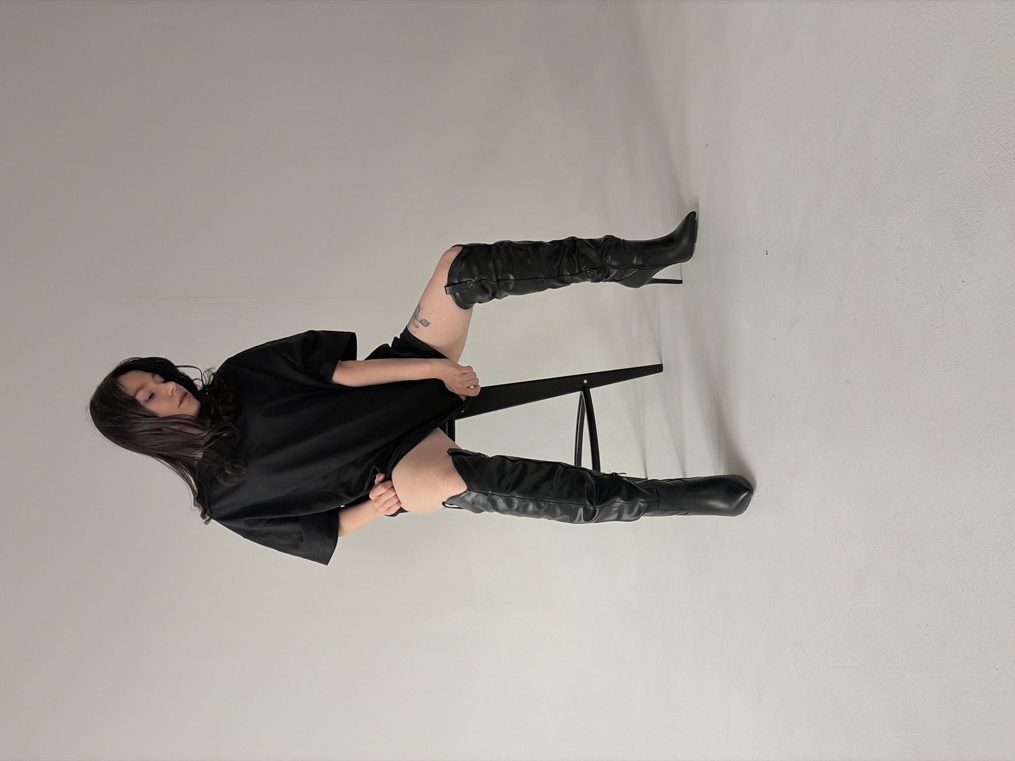 A woman with long, dark hair is sitting on a black chair in a studio setting. She is wearing a black oversized t-shirt and black, knee-high, high-heeled boots. The background is a plain, light grey or white wall. The image appears to be taken indoors in a studio. The woman is posed in a way that her legs are extended, and she is looking downwards. There is a small tattoo on her left thigh. The location is not discernible from the image.