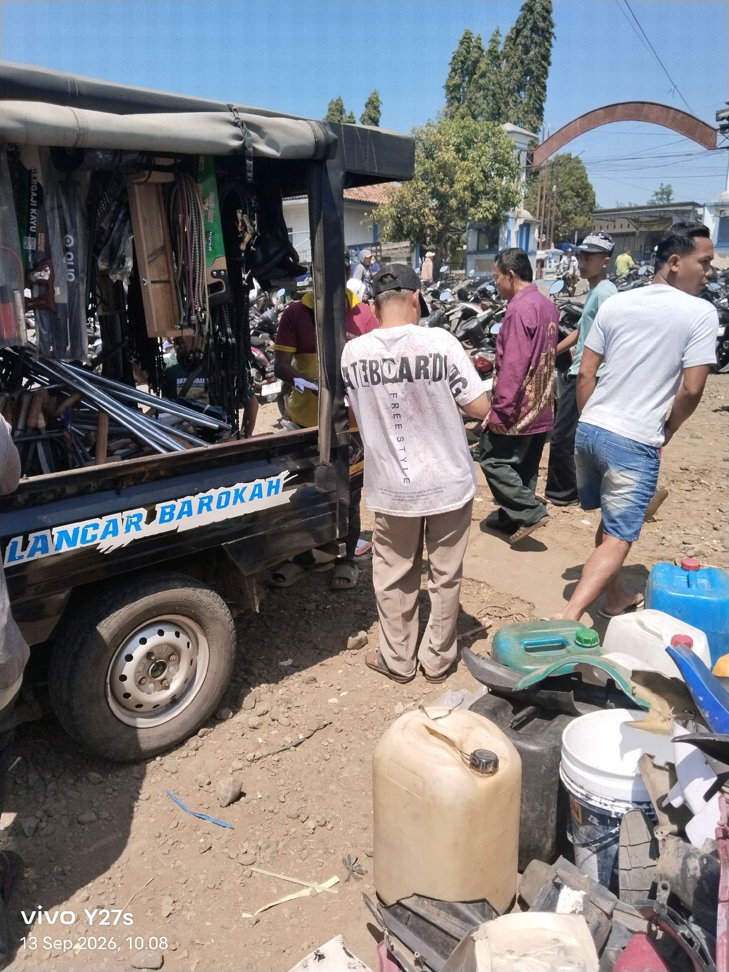 The image depicts an outdoor scene in Trimulyo, Indonesia, on a sunny day. In the foreground, a black truck bed is laden with various items, including what appear to be metal pipes, tools, and other assorted objects. A blue, stylized text graphic reading "LANCAR BAROKAH" is affixed to the side of the truck bed. To the right of the truck, several people are visible. One individual, wearing tan pants and a white t-shirt with "SKATEBOARD" printed on it, stands facing away from the camera. Another person, in a purple patterned shirt and dark pants, is also facing away. A third individual, in a white t-shirt and denim shorts, is seen from the side. Scattered on the ground are several plastic containers, including blue and tan jerrycans, and a white bucket. The ground is covered with loose gravel and dirt. In the background, parked motorcycles are visible, suggesting a market or busy area. A large, arch-shaped structure is present in the distance, along with buildings and trees. The overall impression is a bustling, possibly commercial, outdoor environment.