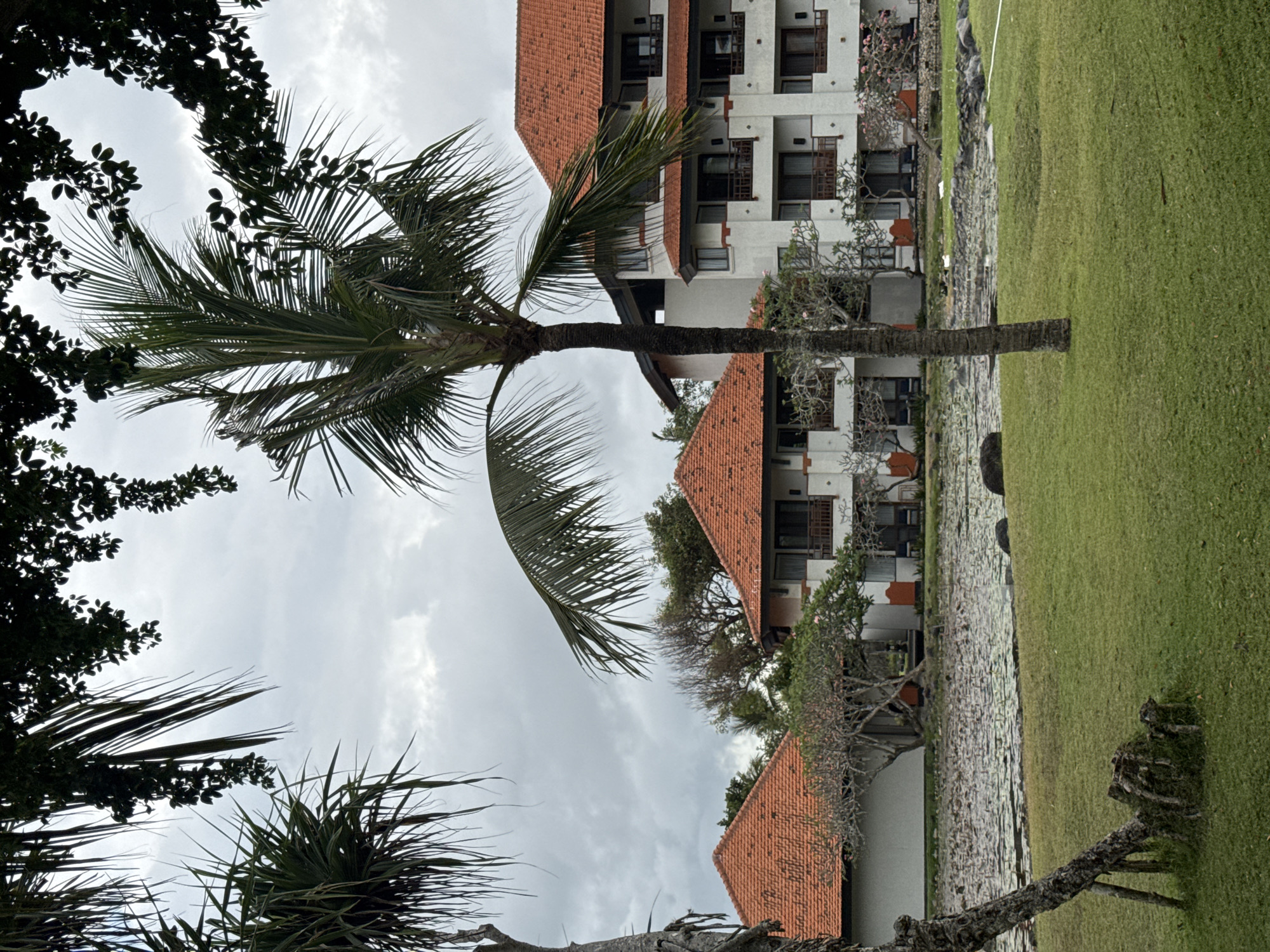 The image shows a resort in Kuta Selatan, Indonesia. The resort features several white buildings with red tile roofs and balconies. In front of the buildings, there is a pond with lily pads and a grassy lawn. Palm trees and other tropical foliage are scattered throughout the scene. The sky is overcast, suggesting it may be a cloudy day or evening. The scene appears peaceful and tranquil, with no people visible in the frame.