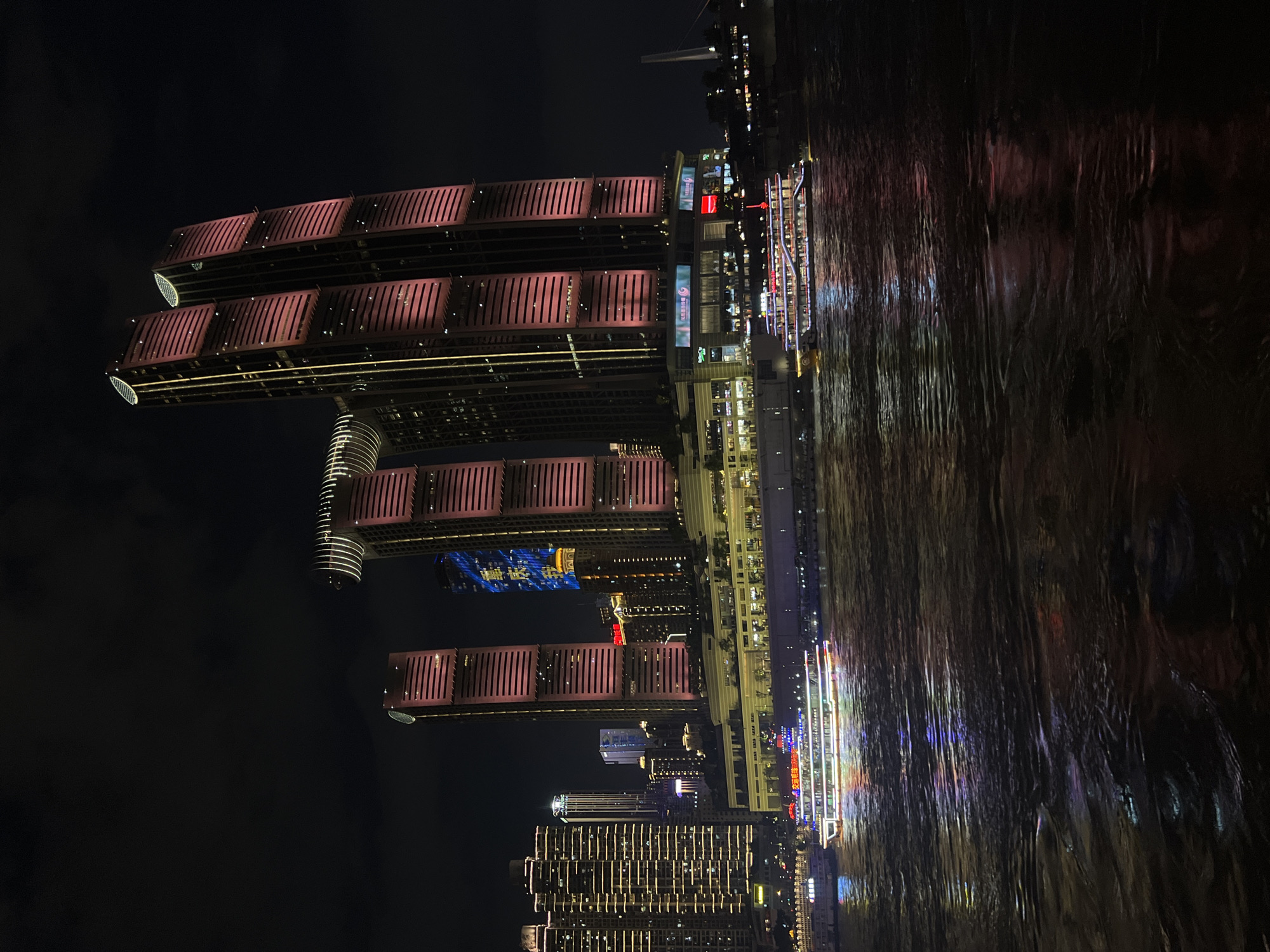 The image depicts a nighttime view of the Yangtze River in Nan'an District, China, with illuminated modern skyscrapers dominating the skyline and a passenger boat on the water. The skyscrapers, notably the Hongyadong complex, are lit with pink and yellow lights, highlighting their architectural features. The water reflects the vibrant lights of the city and the boat. A passenger boat, adorned with colorful lights, is cruising on the river. The scene is a cityscape at night, with the water in the foreground and the buildings in the background. There are no visible people in the image.